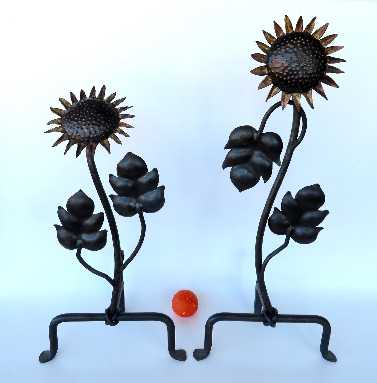 A unique pair of sunflower andirons, custom made in mostly hand forged iron. The sunflowers, each of: A unique pair of sunflower andirons, custom made in mostly hand forged iron. The sunflowers, each of different heights, rising from the base with 2 attached leaves mounted on tri-legs - the front legs