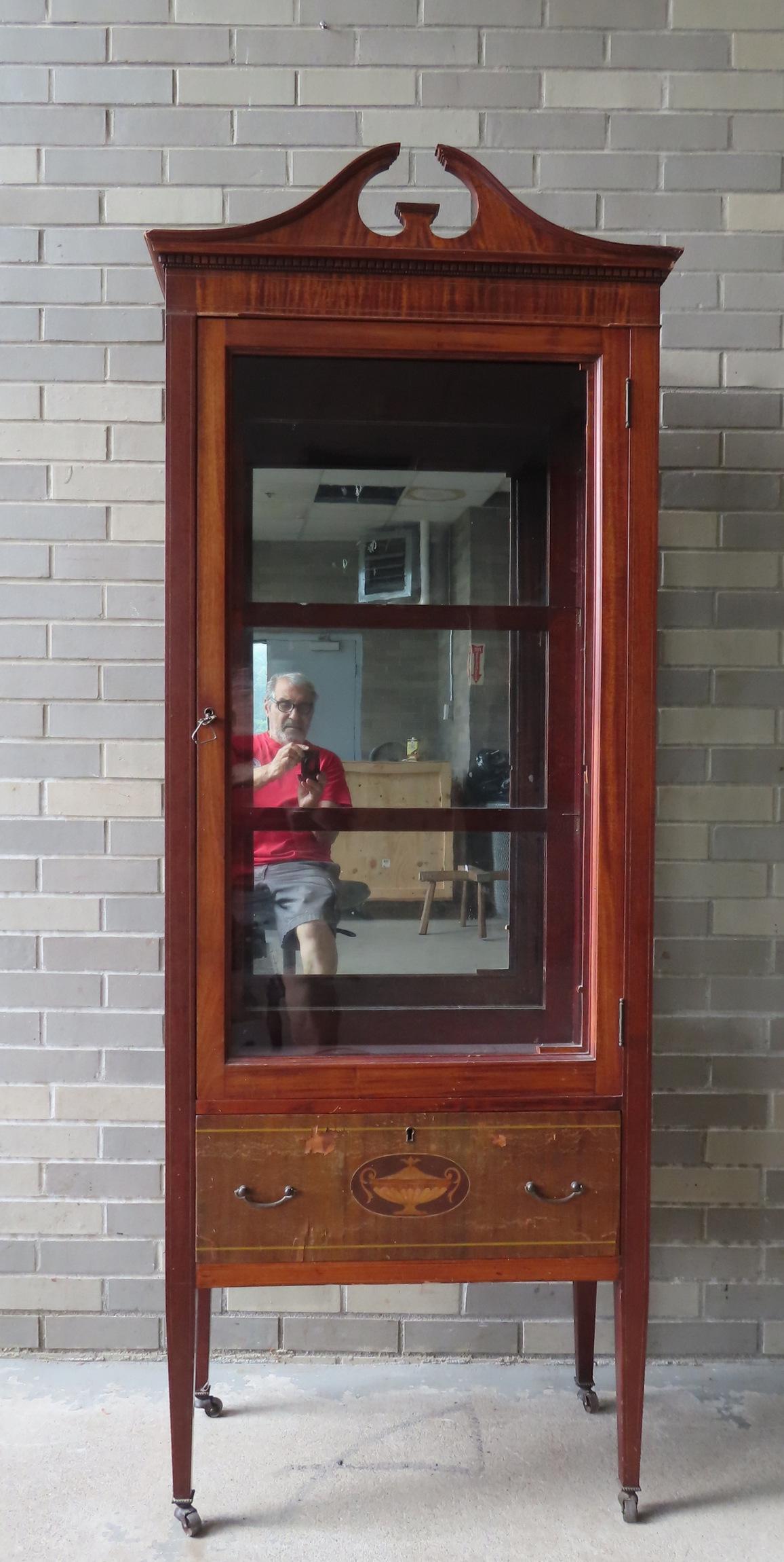 A Hepplewhite style mahogany china cabinet having a broken arch top over 1 door over 1 drawer, circa: A Hepplewhite style mahogany china cabinet having a broken arch top over 1 door over 1 drawer, circa 1900. Line inlays throughout frame and lower drawer with urn inlay. Condition: Missing fretwork mul