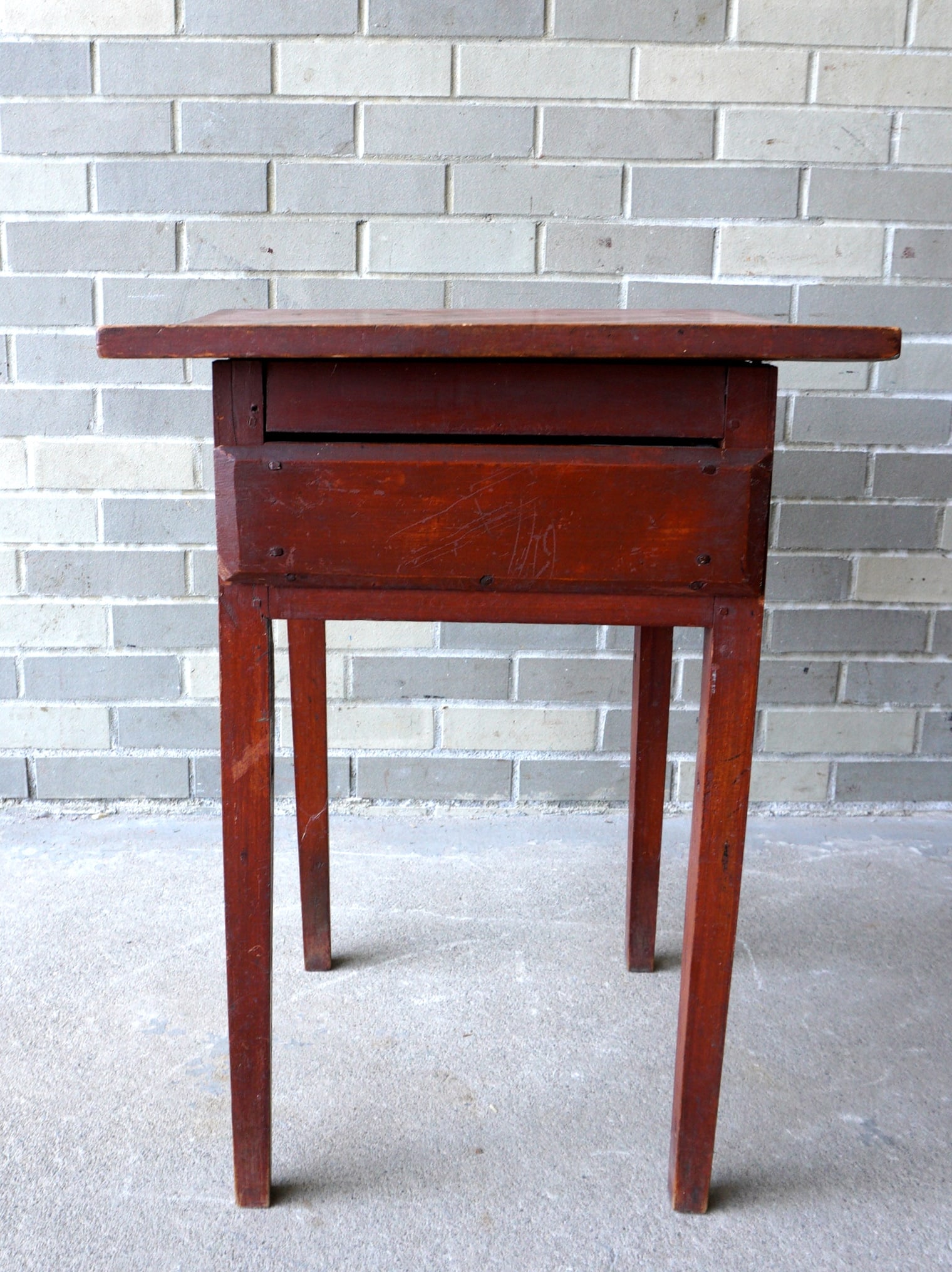 A country Hepplewhite 1 drawer stand of primitive construction in original red stain. The drawer: A country Hepplewhite 1 drawer stand of primitive construction in original red stain. The drawer with chamfered sides is attached to sides but cut nails. One leg with split where it meets the apron, e