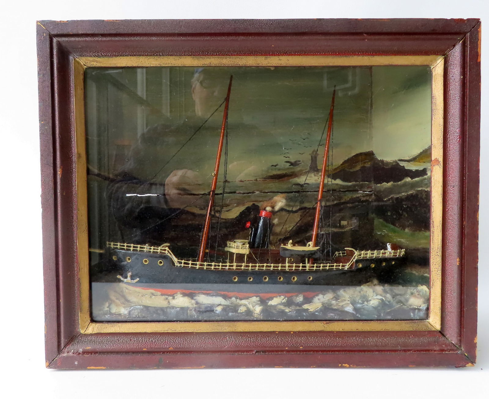 A sailing ship diorama housed in its original shadow box frame with hand painted molded waves and: A sailing ship diorama housed in its original shadow box frame with hand painted molded waves and hand painted landscape in background including lighthouse and sea birds. The ship with stained wool ri