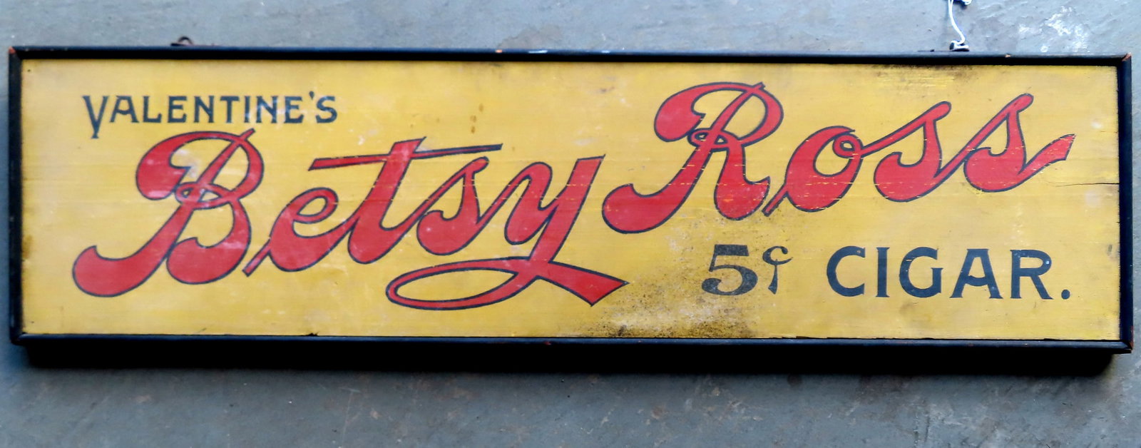 A wooden sign marked "Valentine's Betsy Ross 5 cents Cigars". The sign with red and black lettering: A wooden sign marked "Valentine's Betsy Ross 5 cents Cigars". The sign with red and black lettering on yellow ground and with framed edges. It does show minor wear and some discoloration but difficult