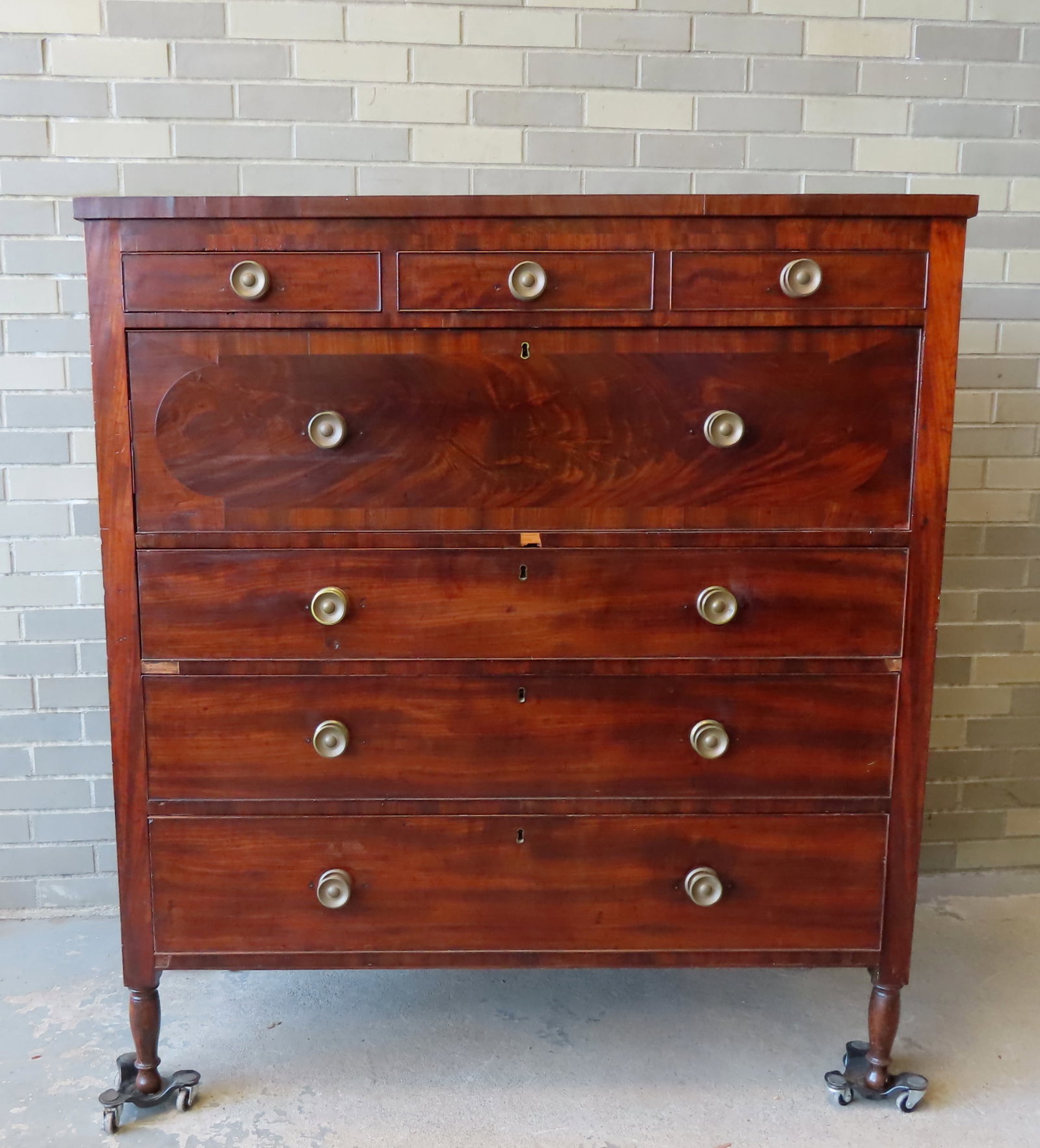 A Sheraton mahogany chest of drawers having 3 short drawers over 1 large drawer with a shaped: A Sheraton mahogany chest of drawers having 3 short drawers over 1 large drawer with a shaped mahogany inlaid panel over 3 graduated drawers - mounted on turned feet. The brass pulls, while matching, 