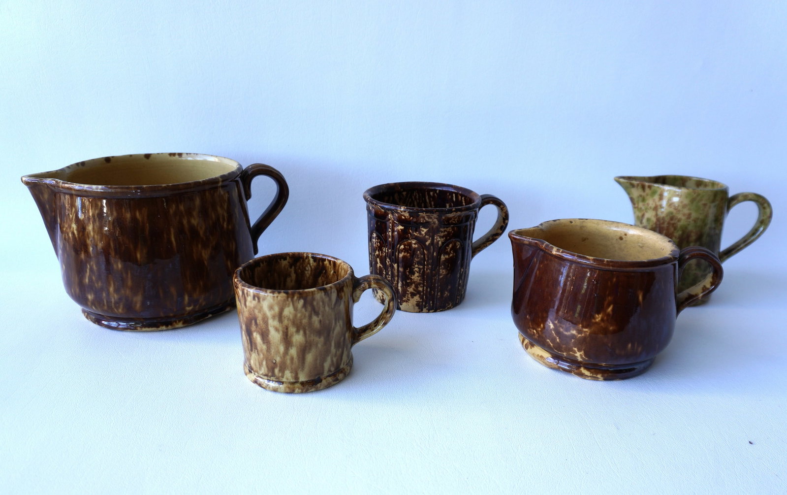 Five assorted Bennington pitchers and cups, all 19th century. The largest, with hairline in base,: Five assorted Bennington pitchers and cups, all 19th century. The largest, with hairline in base, measures 9"W x 5"H. Two cups with hairlines in base. The pitcher with green and mustard color is in ve