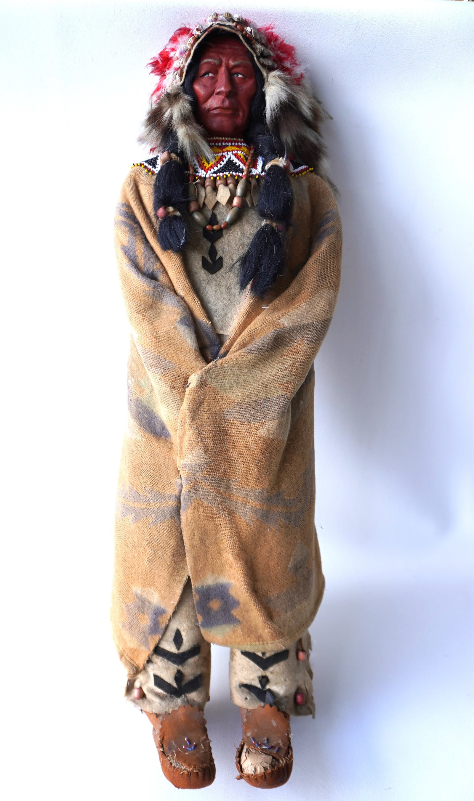A large country store Skookum doll display in original costume - approximately 36"L x 12"W. Losses: A large country store Skookum doll display in original costume - approximately 36"L x 12"W. Losses to feathers on bonnet, blanket with faded colors, and losses to moccasins. Early to mid 20th century.