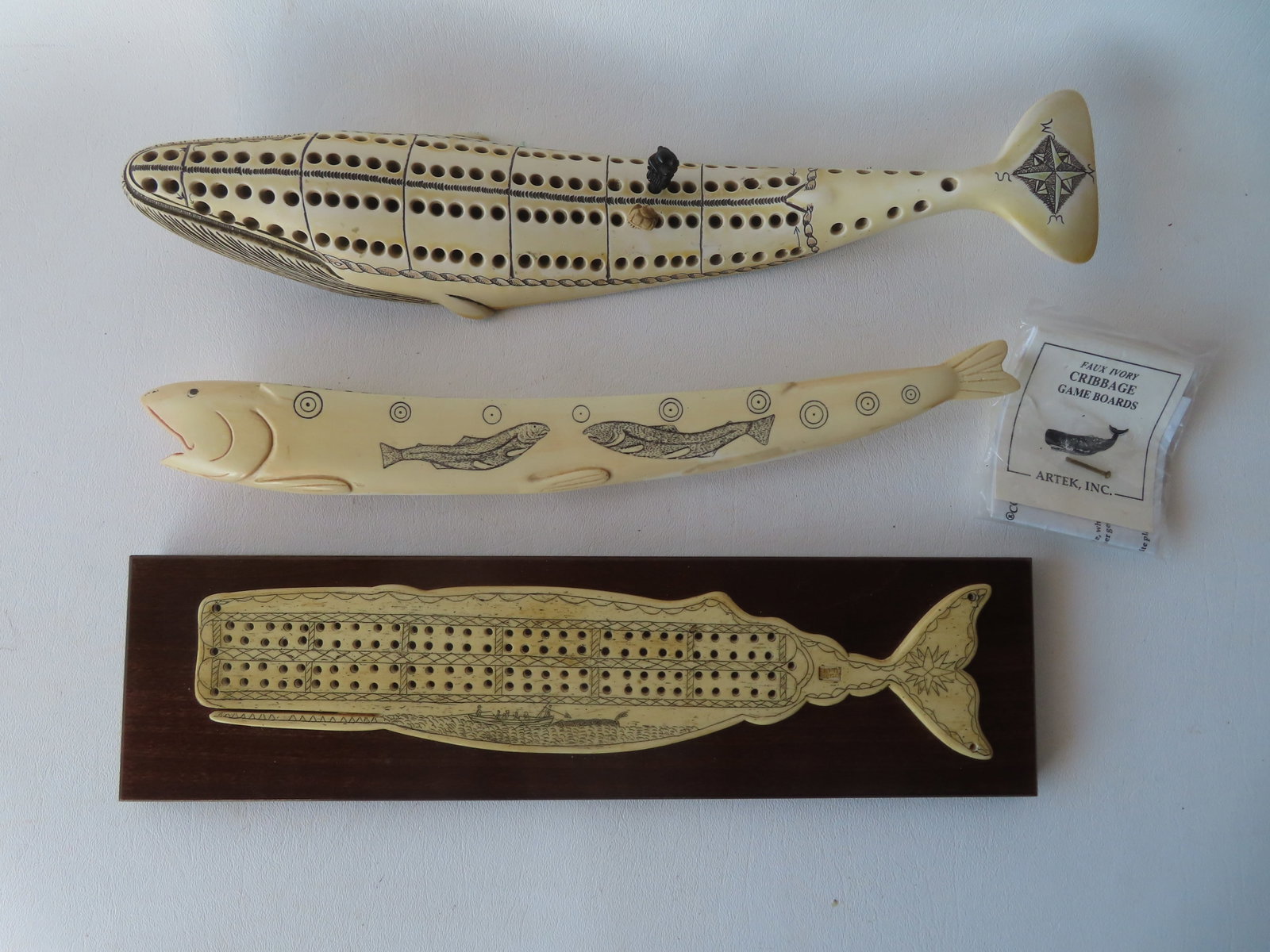 Three resin whale tooth scrimshaw cribbage boards. The first, in the form of a fish signed "KWM: Three resin whale tooth scrimshaw cribbage boards. The first, in the form of a fish signed "KWM Artek 1992" - very good condition - 12 1/2"L; the second of a swimming whale - 13 1/2"L - very good cond