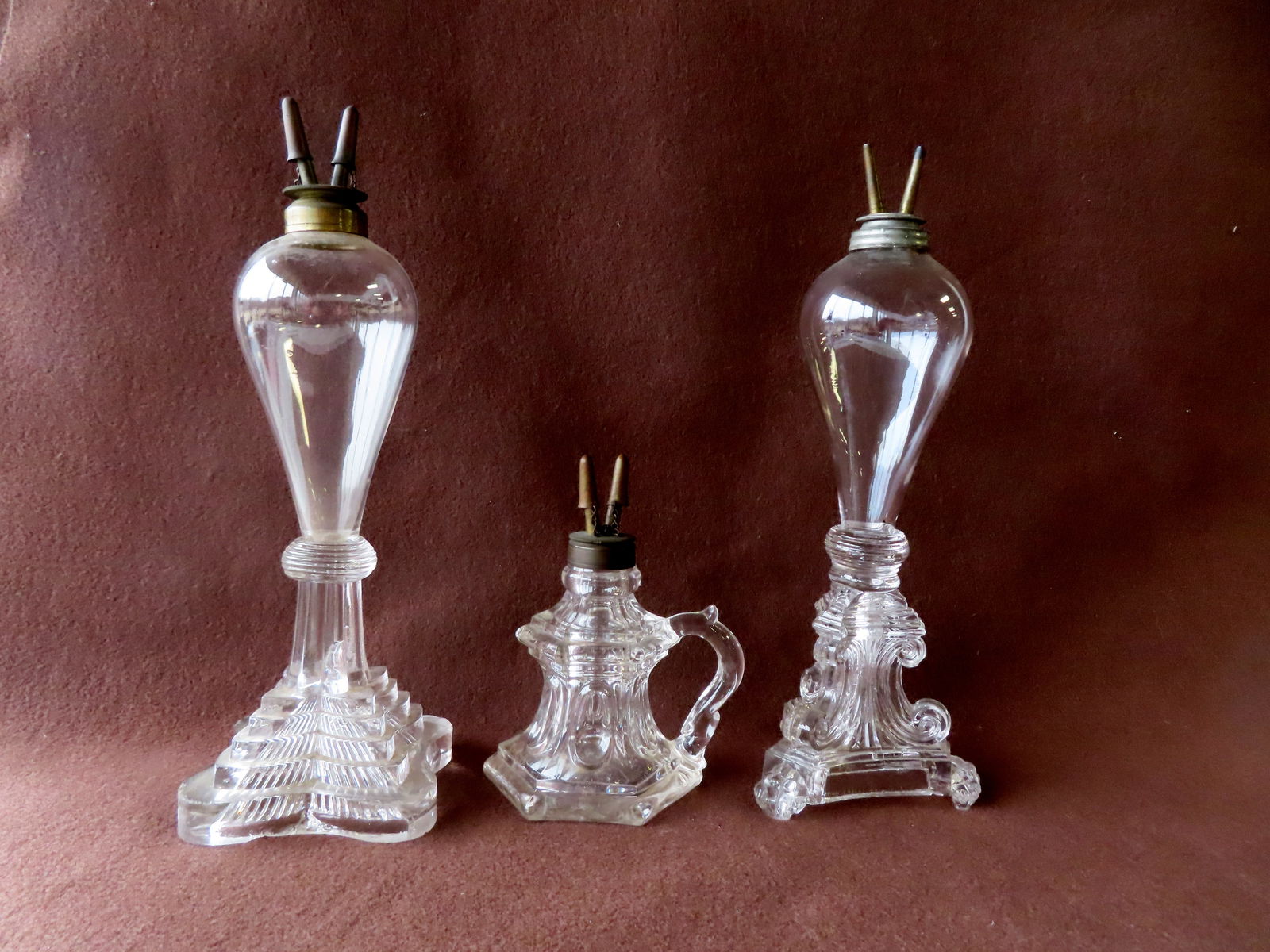 Three colorless whale oil lamps with burners, one a finger lamp with pontil mark on base in very: Three colorless whale oil lamps with burners, one a finger lamp with pontil mark on base in very good condition, and 2 appearing to be Sandwich with blown bulb shaped fonts and pressed bases. One base