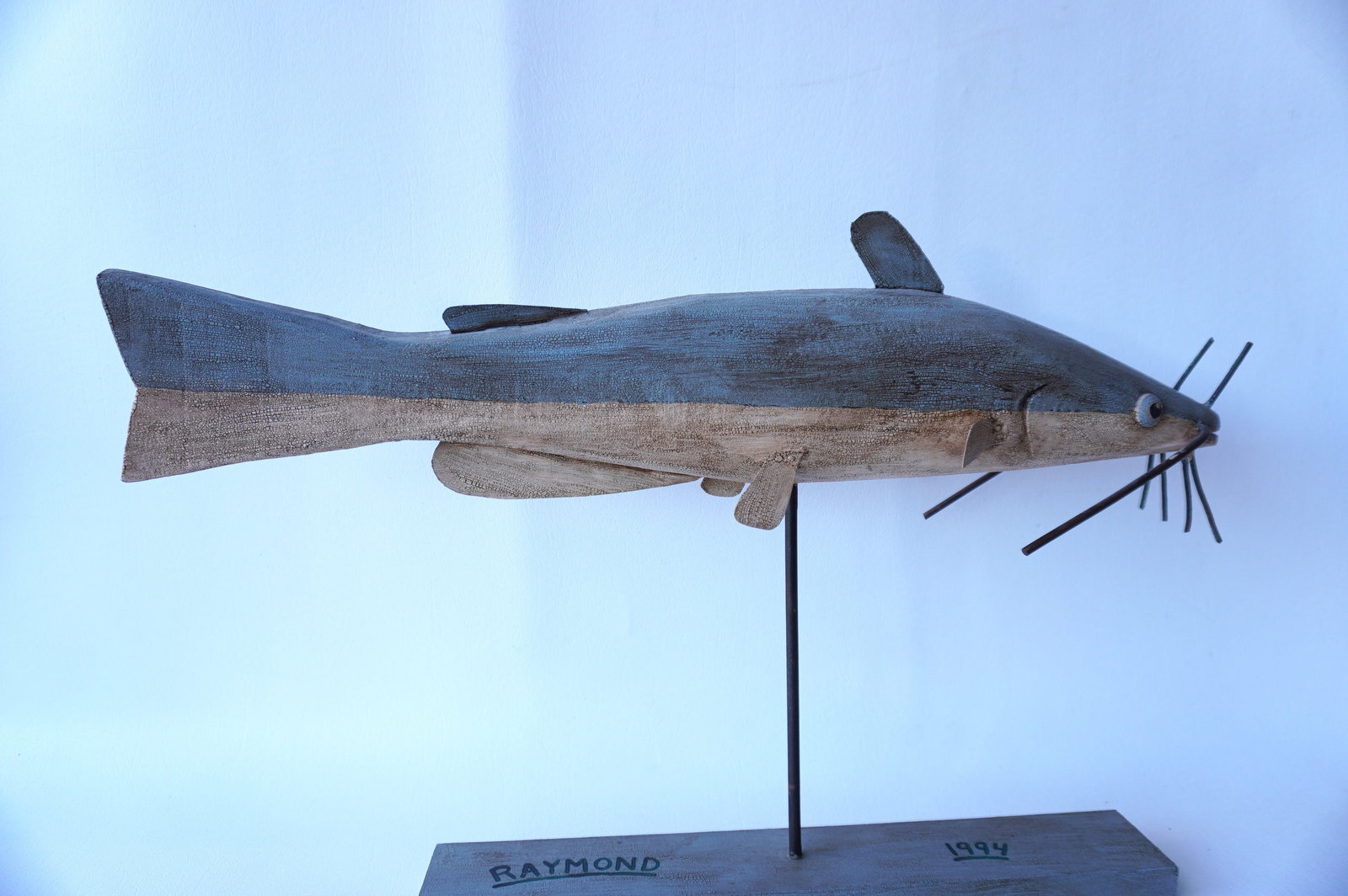 Jeff Raymond, folk art fish carving (1949-2022, Poughkeepsie and Red Hook NY). A stylized wooden: Jeff Raymond, folk art fish carving (1949-2022, Poughkeepsie and Red Hook NY). A stylized wooden Catfish carving in original paint with tin fins and mounted on wooden platform - signed on base Raymond