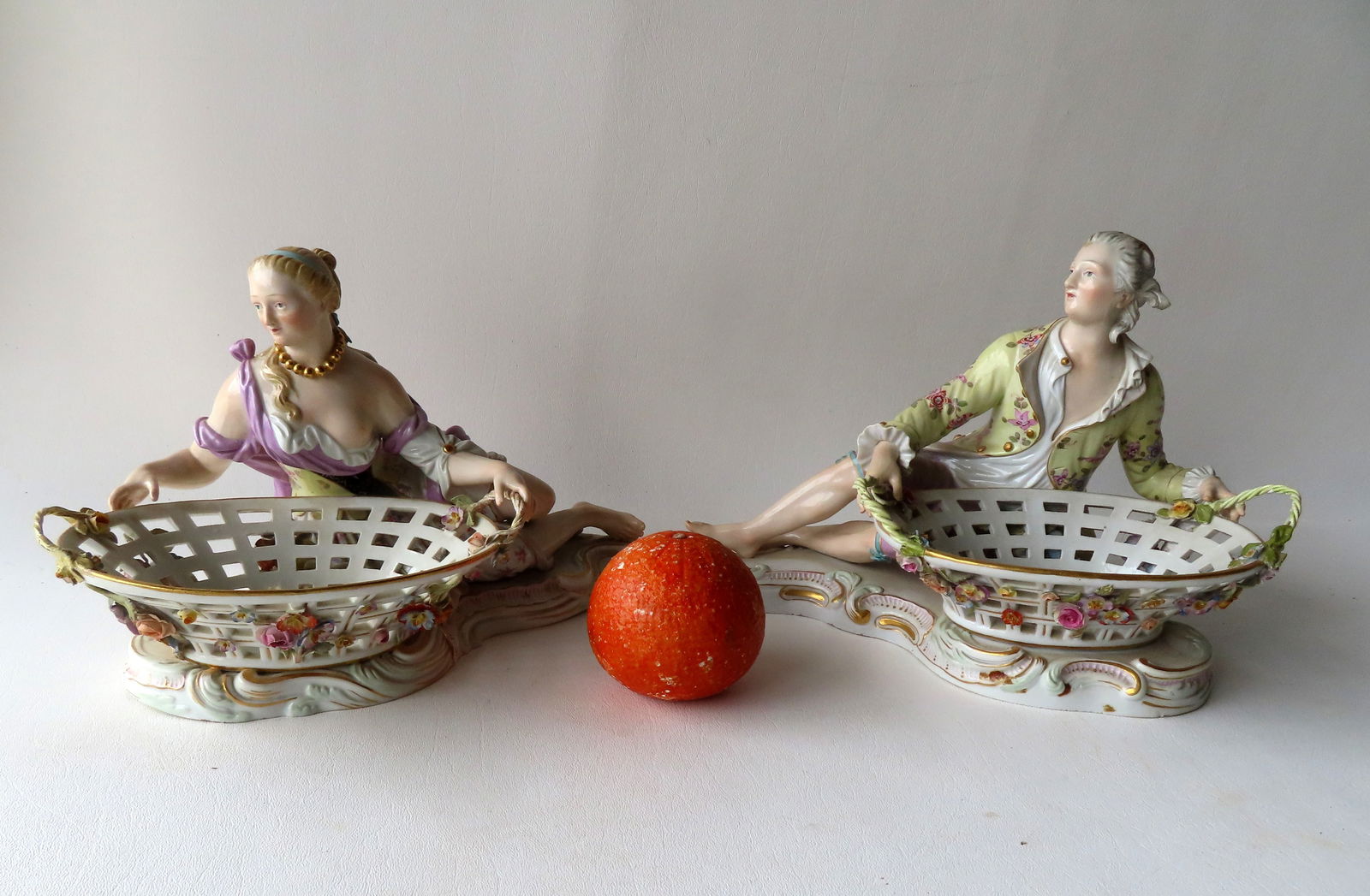 A pair of matching Meissen porcelain reclining figures of man and woman with open weave baskets,: A pair of matching Meissen porcelain reclining figures of man and woman with open weave baskets, likely for holding bonbons, a popular chocolate of the time, late 19th to early century. They both have