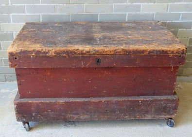 A primitive tool box in original red paint, mounted on wheels, recessed iron handles, and having
