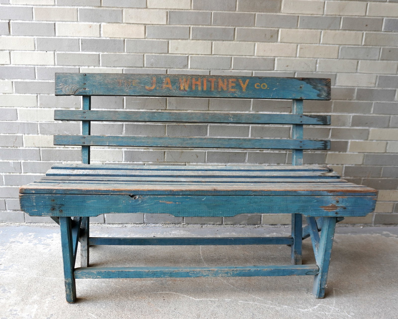 A slat back wooden park bench in original blue/green paint and signed in yellow lettering across the: A slat back wooden park bench in original blue/green paint and signed in yellow lettering across the top back slat "The J.A. Whitney & Co.". Paint wear and losses, slight wobble to right and left, oth