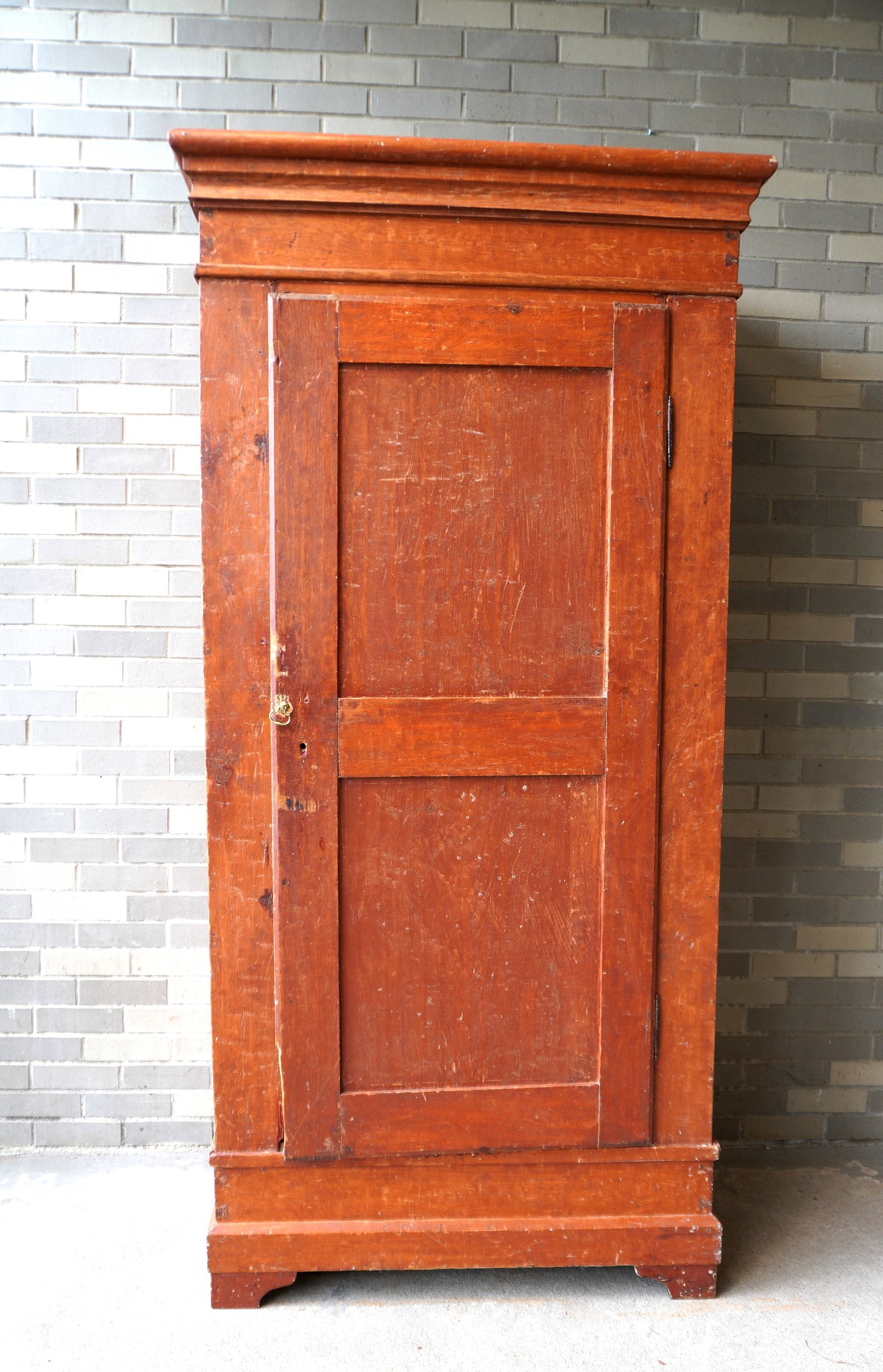 A primitive country cupboard/armoire, singled paneled door, scraped down to original salmon red: A primitive country cupboard/armoire, singled paneled door, scraped down to original salmon red stain, having 1 shaped shelf over a newer wooden clothes hanger. The cupboard with its original cornice 