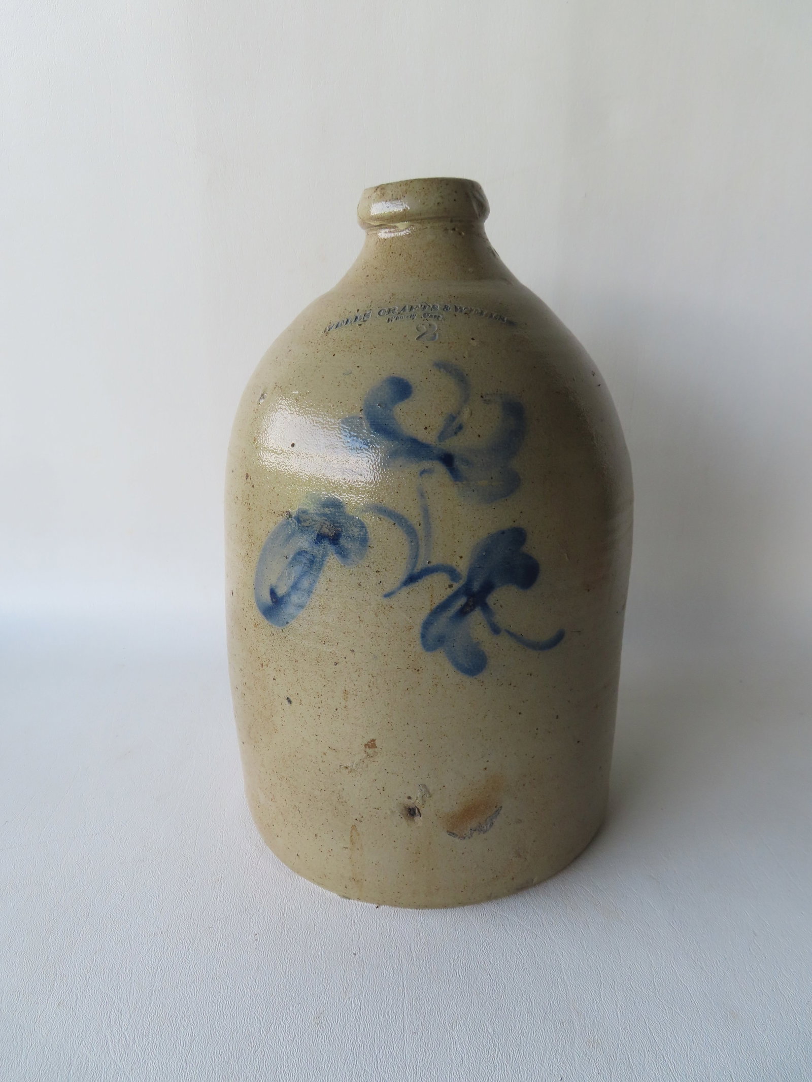 A stoneware jug decorated with a cobalt blue flower with the impressed mark of "WELLS CRAFTS & WELLS: A stoneware jug decorated with a cobalt blue flower with the impressed mark of "WELLS CRAFTS & WELLS / Whately Mass.", 2 gallon size. Tight hairlines on handle and lip, firing burst on body, else good
