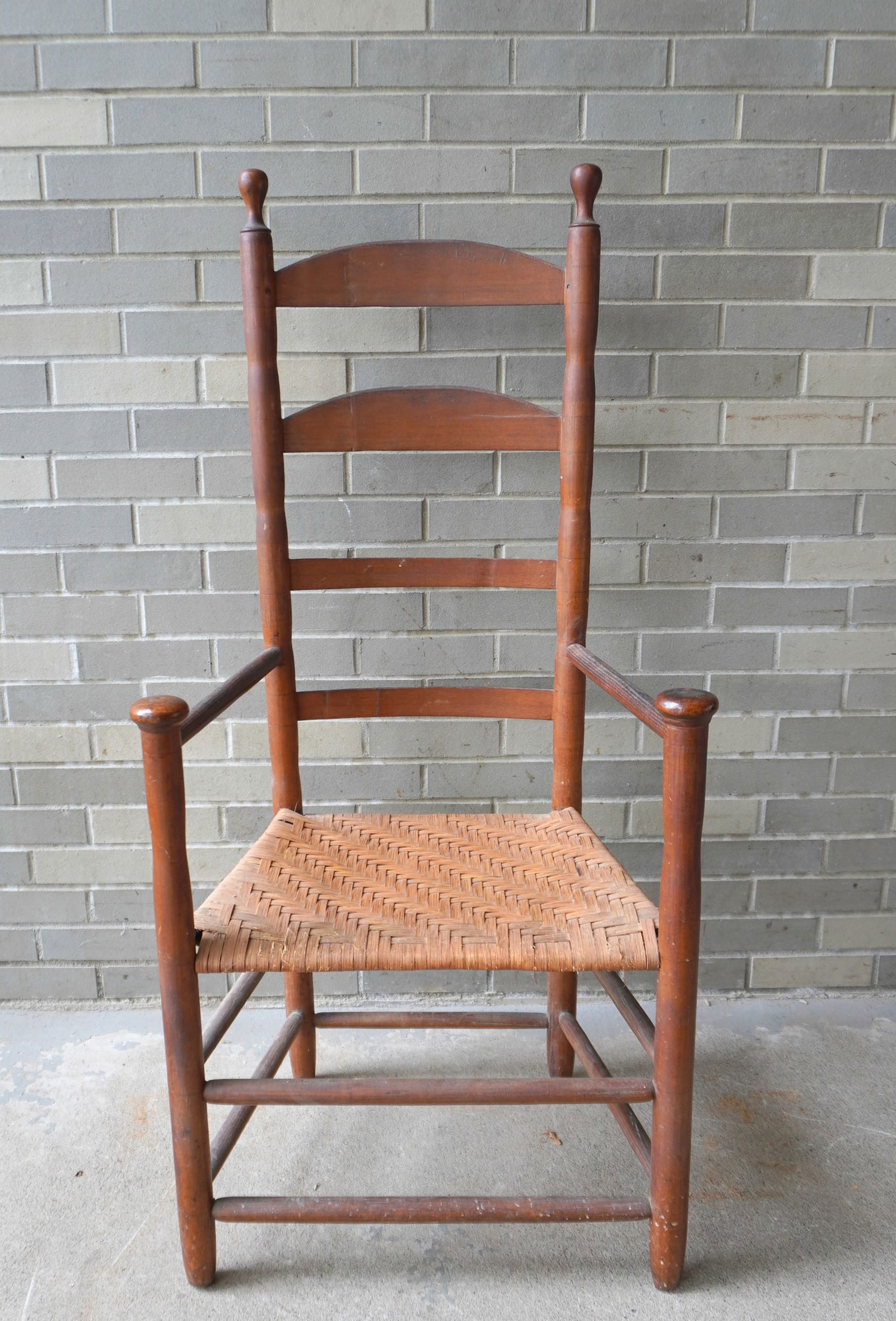 Ladder back arm chair with mushroom finials on arms, early 19th century in natural finish. Replaced: Ladder back arm chair with mushroom finials on arms, early 19th century in natural finish. Replaced woven cane seat otherwise good condition. Measures 21 1/2"H x 16"D x 49 1/4"H. Provenance: From an e