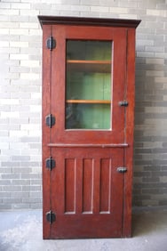 A primitive pine cupboard in original red stain, single glass door over a paneled door and having a