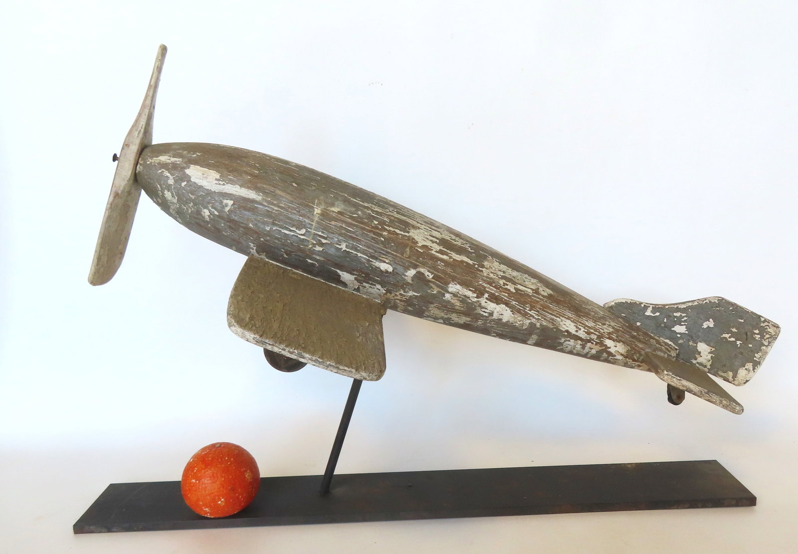 A folk art carved wooden airplane weathervane with single wing with applied wheels and having a: A folk art carved wooden airplane weathervane with single wing with applied wheels and having a wooden propeller attached to nose - old white paint over traces of original gray paint and in good condi