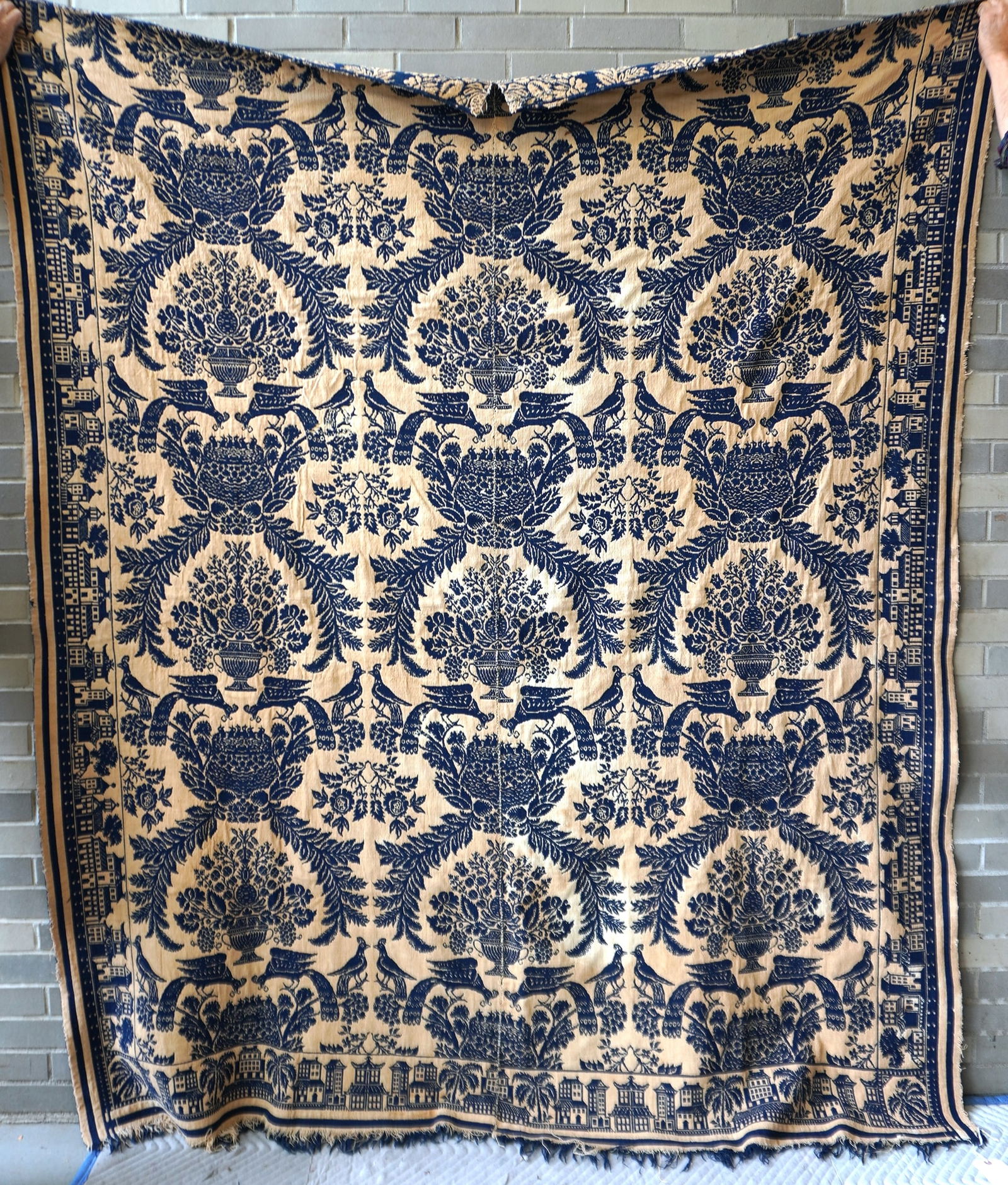 A Jacquard blue and white coverlet having a border decorated with a series of buildings and trees: A Jacquard blue and white coverlet having a border decorated with a series of buildings and trees and a central field of alternating medallions of flowers in urns and pheasants feeding nestlings - the