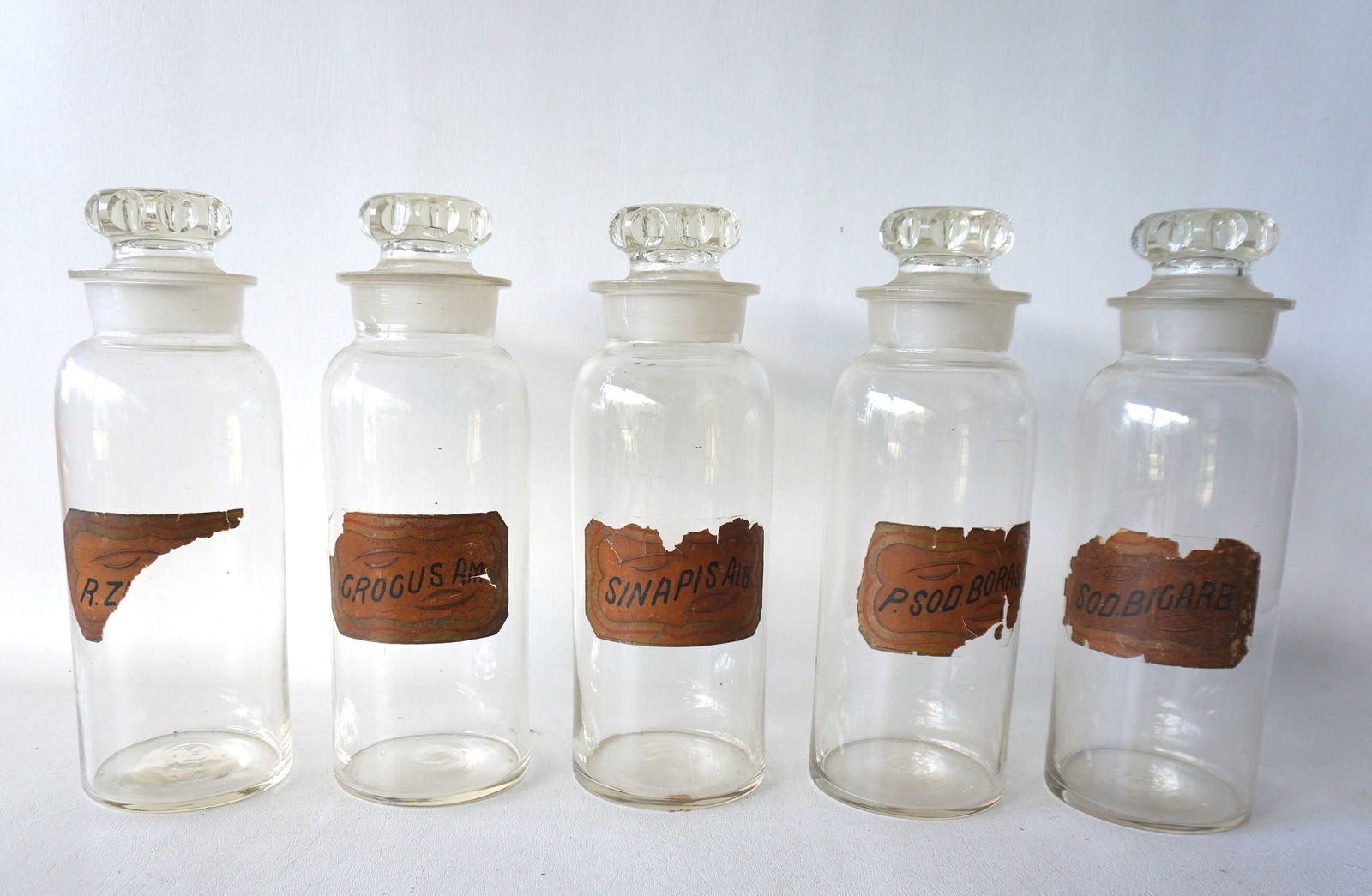 Grouping of 5 large apothecary jars with matching stoppers and with original paper labels still: Grouping of 5 large apothecary jars with matching stoppers and with original paper labels still attached, late 19th century. All of the attached paper labels are with losses, but the jars and stoppers