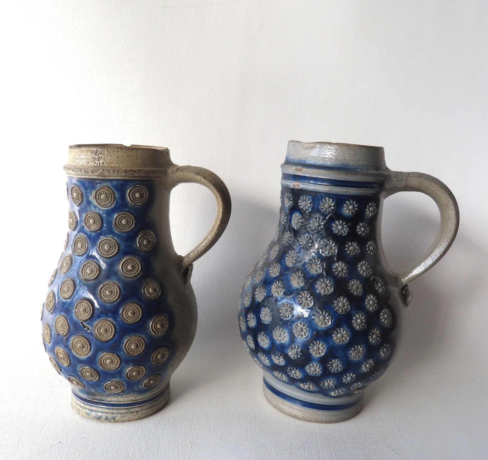 Two Westerwald stoneware tankards, lacking lids, both in brushed cobalt blue and having embossed: Two Westerwald stoneware tankards, lacking lids, both in brushed cobalt blue and having embossed floral button on the body. One with several small rim chimps and the other with 3 rim chip repairs, oth