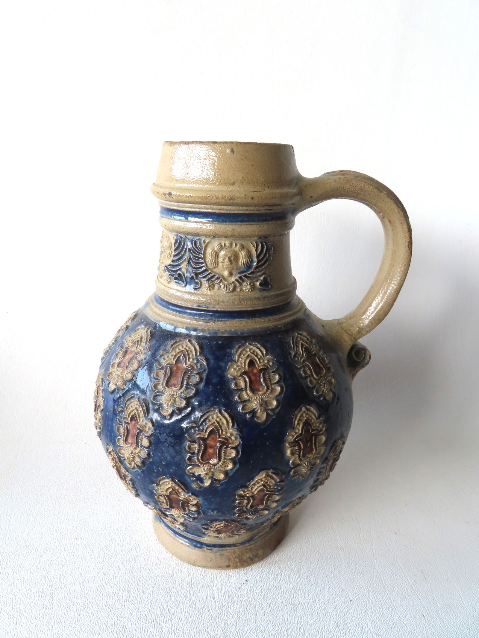 A Westerwald bulbous stoneware jug, decorated with medallions around the body with central images of: A Westerwald bulbous stoneware jug, decorated with medallions around the body with central images of a flower that are brushed in plum manganese, the background brushed in cobalt blue - the neck decor
