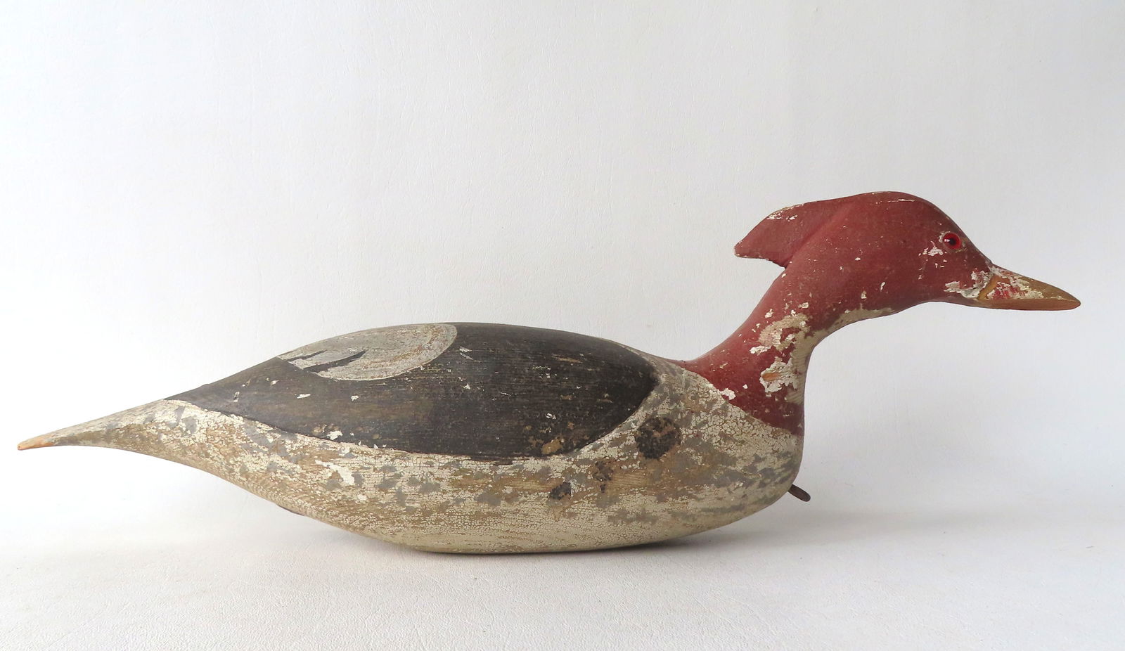 A swimming Merganser decoy, weathered original paint, glass eyes, carved wings, and offset lead: A swimming Merganser decoy, weathered original paint, glass eyes, carved wings, and offset lead weight in base - loss to underside of beak, some paint wear and losses, else good condition. Measures 18