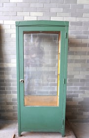 A tall sheet iron medical cabinet with 1 long glass door and glass sides with a pitched roof and 2