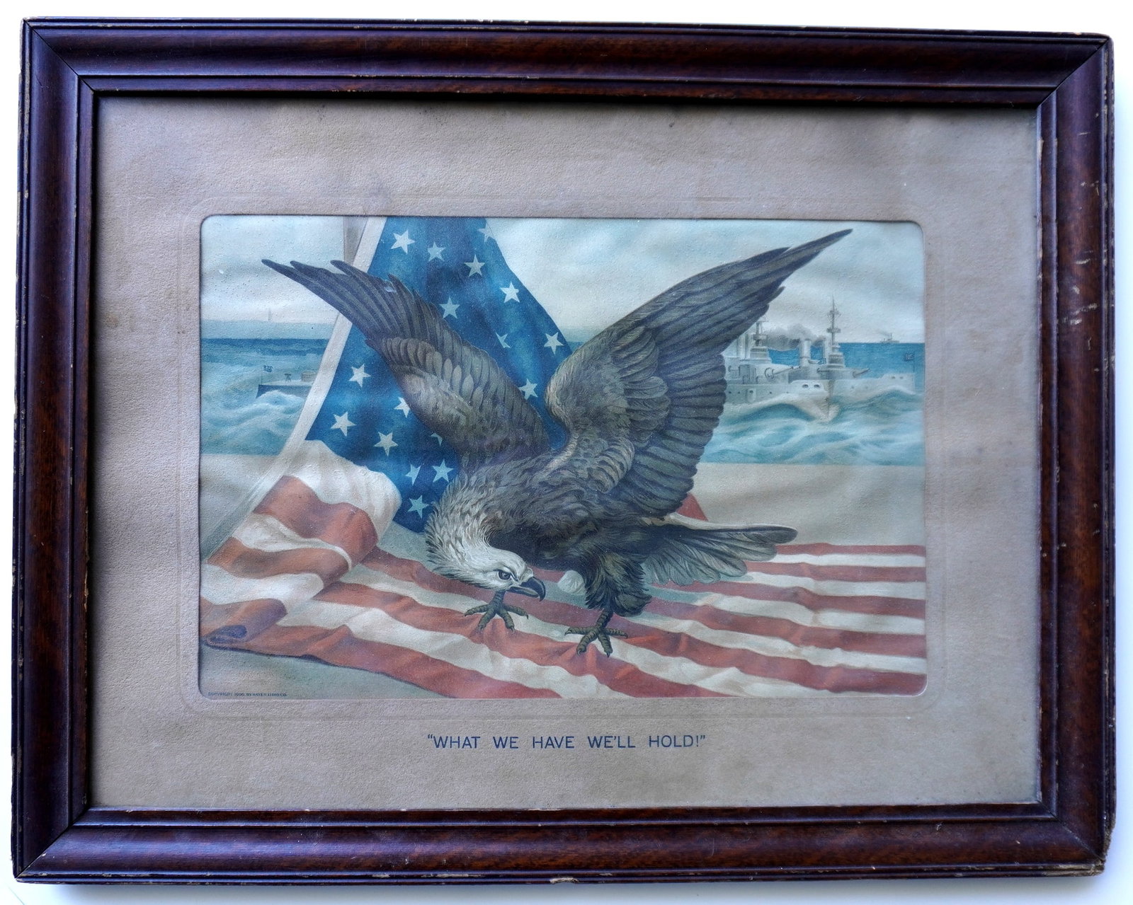 A chromolithograph of a spread wing eagle with American flag in foreground and battleship in: A chromolithograph of a spread wing eagle with American flag in foreground and battleship in background, signed "Copyright 1900 Hays Litho Co." and titled on mat "What We Have We'll Hold" - probably a