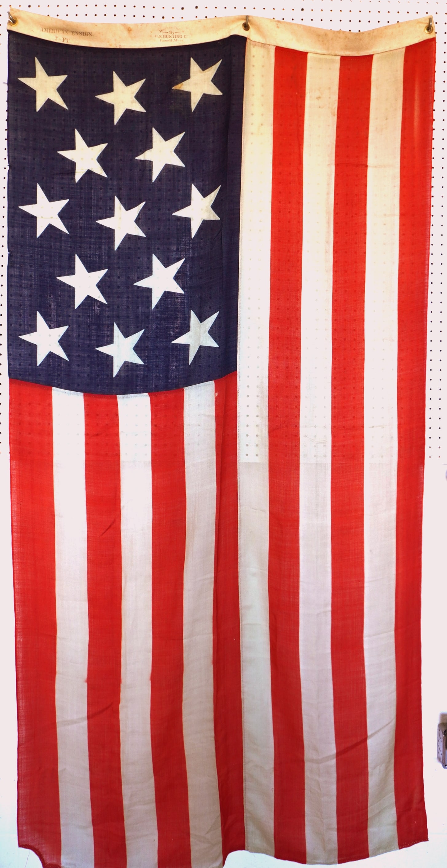 An original Centennial 13 star naval flag signed "U.S. Bunting Co. / Lowell, Mass.". It is also: An original Centennial 13 star naval flag signed "U.S. Bunting Co. / Lowell, Mass.". It is also marked "American Ensign / 7 FT.". This is a press-dyed (printed) flag having 3 sections that are hand se