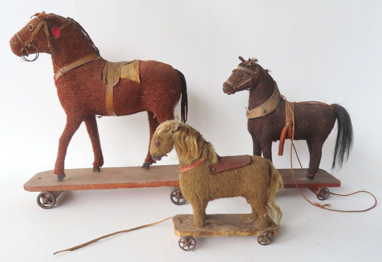 Three primitive horse pull toys mounted on platforms with tin wheels. All missing saddles and: Three primitive horse pull toys mounted on platforms with tin wheels. All missing saddles and harnesses, 2 missing 1 tack eye, and typical use wear, 19th century. The largest measures 12"L x 3 3/4"W x