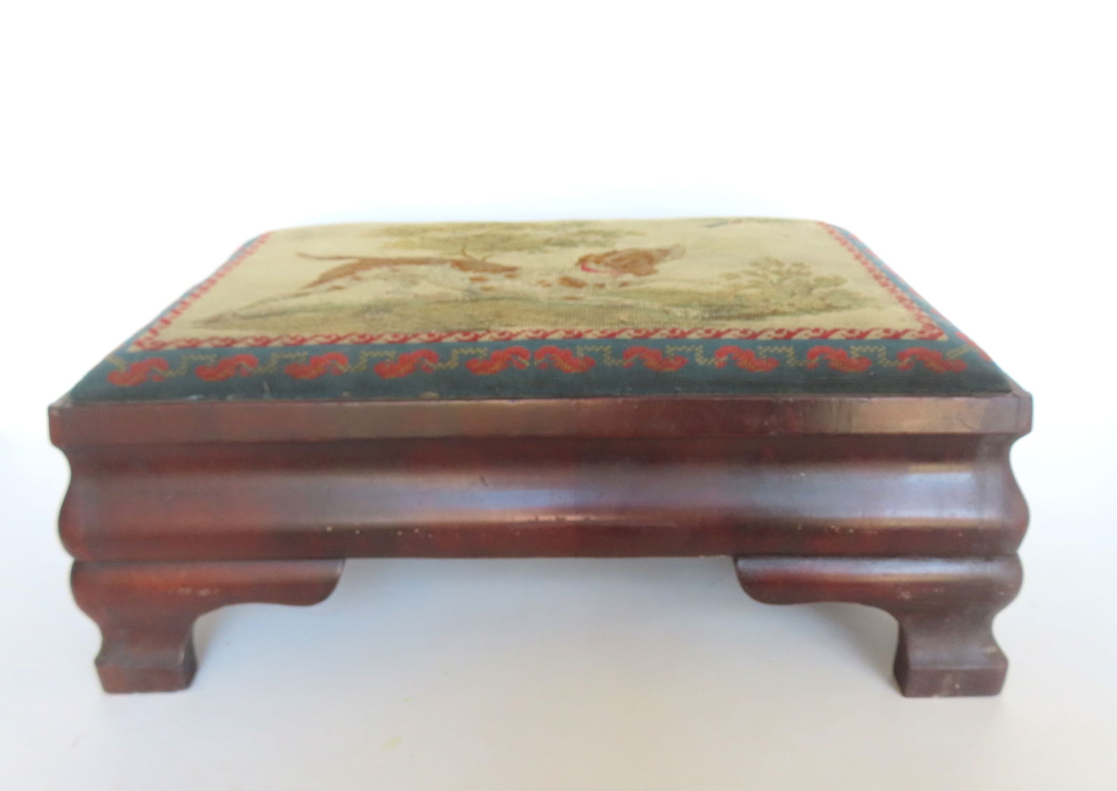 An Empire footstool with needlepoint top depicting a sporting dog chasing a flying bird. The cushion: An Empire footstool with needlepoint top depicting a sporting dog chasing a flying bird. The cushion mounted on a mahogany ogee bracket base. It is in very good condition circa 1850. Measures 25 1/2"W