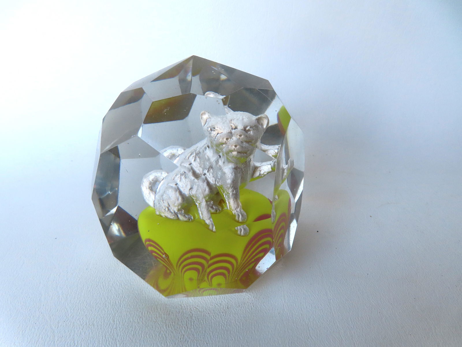 A multi-faceted art glass paperweight encasing a cat holding ball in paw and seated on green: A multi-faceted art glass paperweight encasing a cat holding ball in paw and seated on green cushion, probably mid century. Several tiny chips on corners of facet hardly noticeable and expected use we