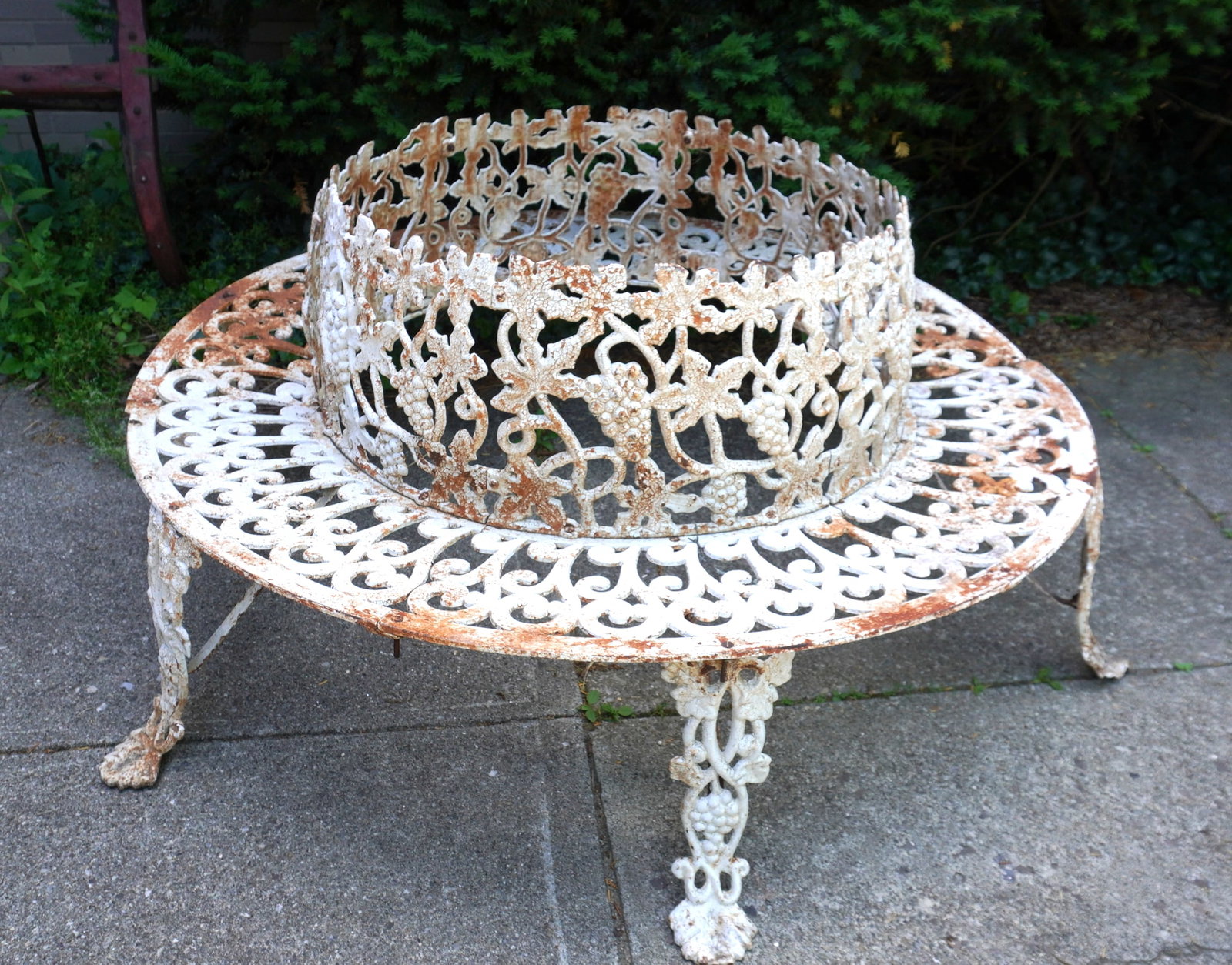 A circular cast iron garden bench in the grape and leaf pattern, designed to fit around a tree, mid: A circular cast iron garden bench in the grape and leaf pattern, designed to fit around a tree, mid 20th century. Missing one bolt that attaches a leg to the, otherwise in good condition. The opening 