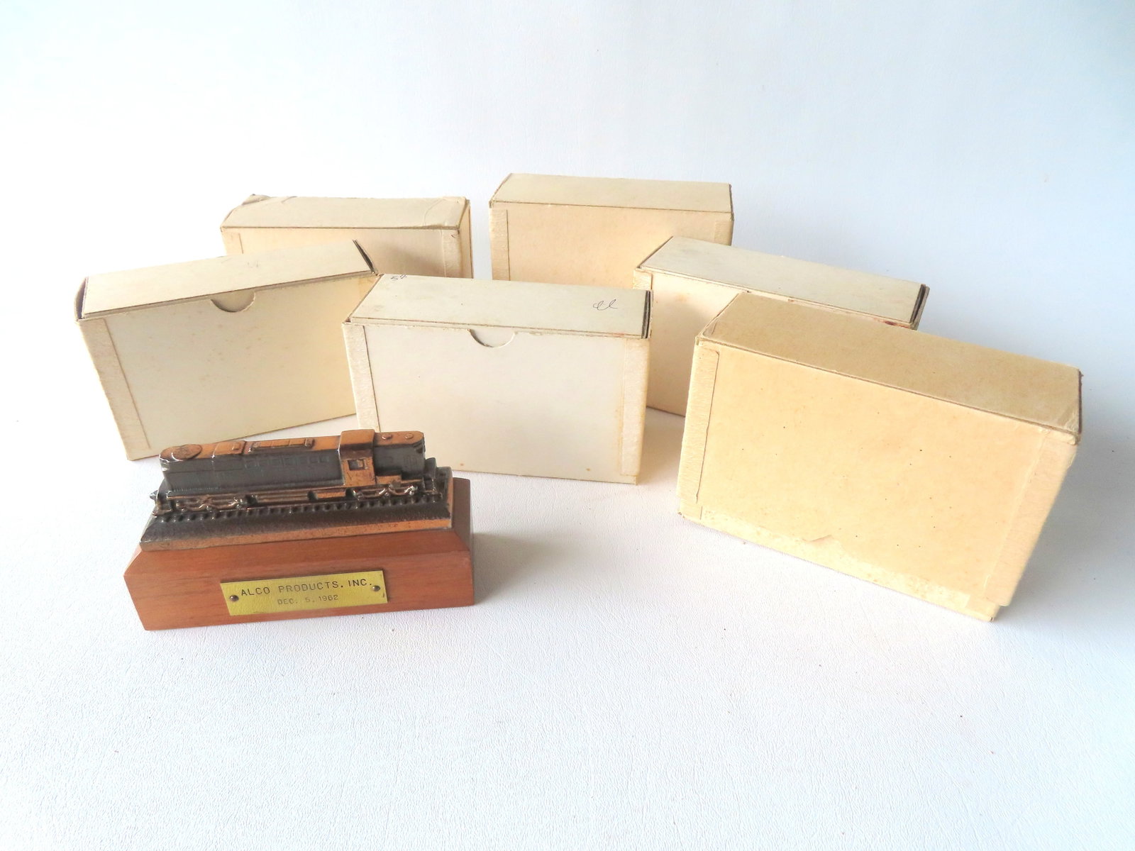 Grouping of 6 unused boxed presentation paper weights in the form of a locomotive, and 1 unboxed,: Grouping of 6 unused boxed presentation paper weights in the form of a locomotive, and 1 unboxed, all in faux bronze finish and marked on brass plaque attached to shaped wooden base "ALCO PRODUCTS INC