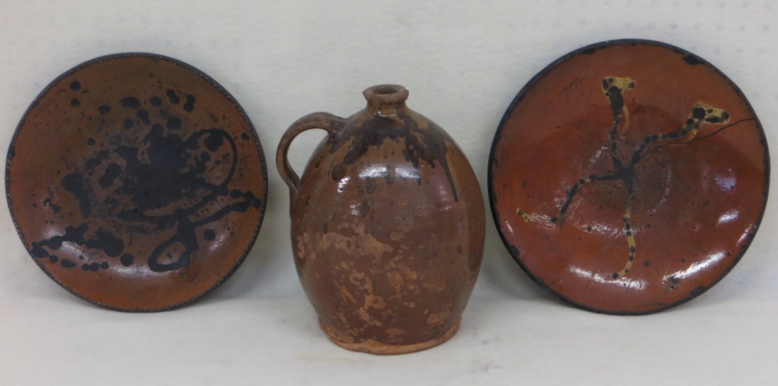 Three pieces of redware, all with some damage including: Three pieces of redware, all with some damage including 2 plates with glaze losses and crack, one with yellow slip decoration (11.75"D & 12.5"D) and a jug with manganese decorations with roughness aro