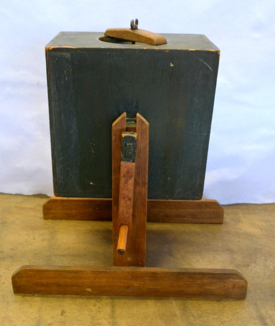 Crank activated revolving butter churn on stand in old: Crank activated revolving butter churn on stand in old green paint. The churn is turned by a handle which causes it to revolve on a wooden axel attached to separate matching stand with long feet. The
