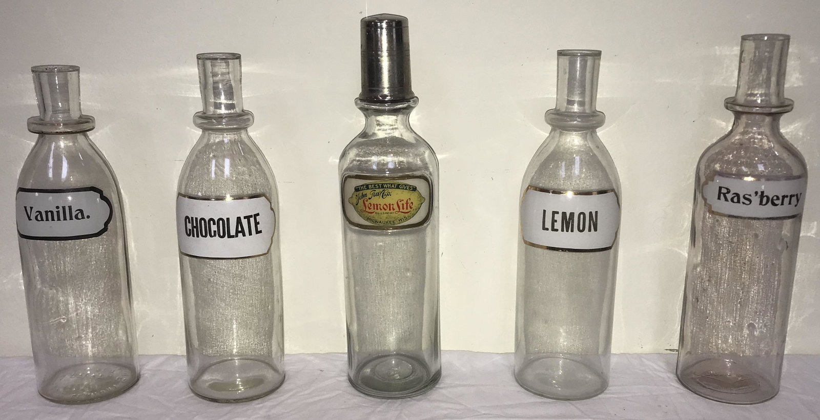 Vintage Clearn Glass Soda Fountain Syrup Bottles: (5) vintage clear glass soda fountain syrup bottles: Lemon Life by John Graf Co., Milkwaukee, WIS., w/silver plated cap, 12 1/2" T x 3 1/4" base diameter; Ras'berry w/slight purple tint in glass, 11 7