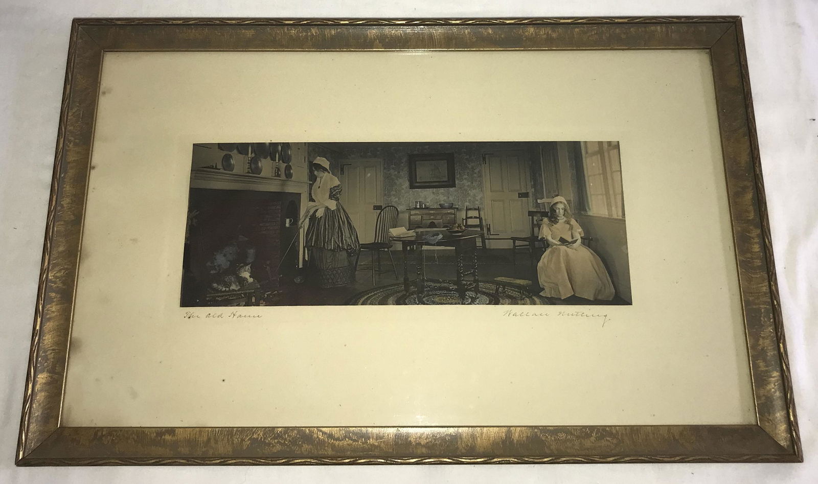 Wallace Nutting Handcolored Photograph: Wallace Nutting Handcolored Photograph, interior scene of 2 woman in a room (1 reading by the window and stoking the fire entitled "The Old House", signed l.r. Wallace Nutting, mounted in a gold tone