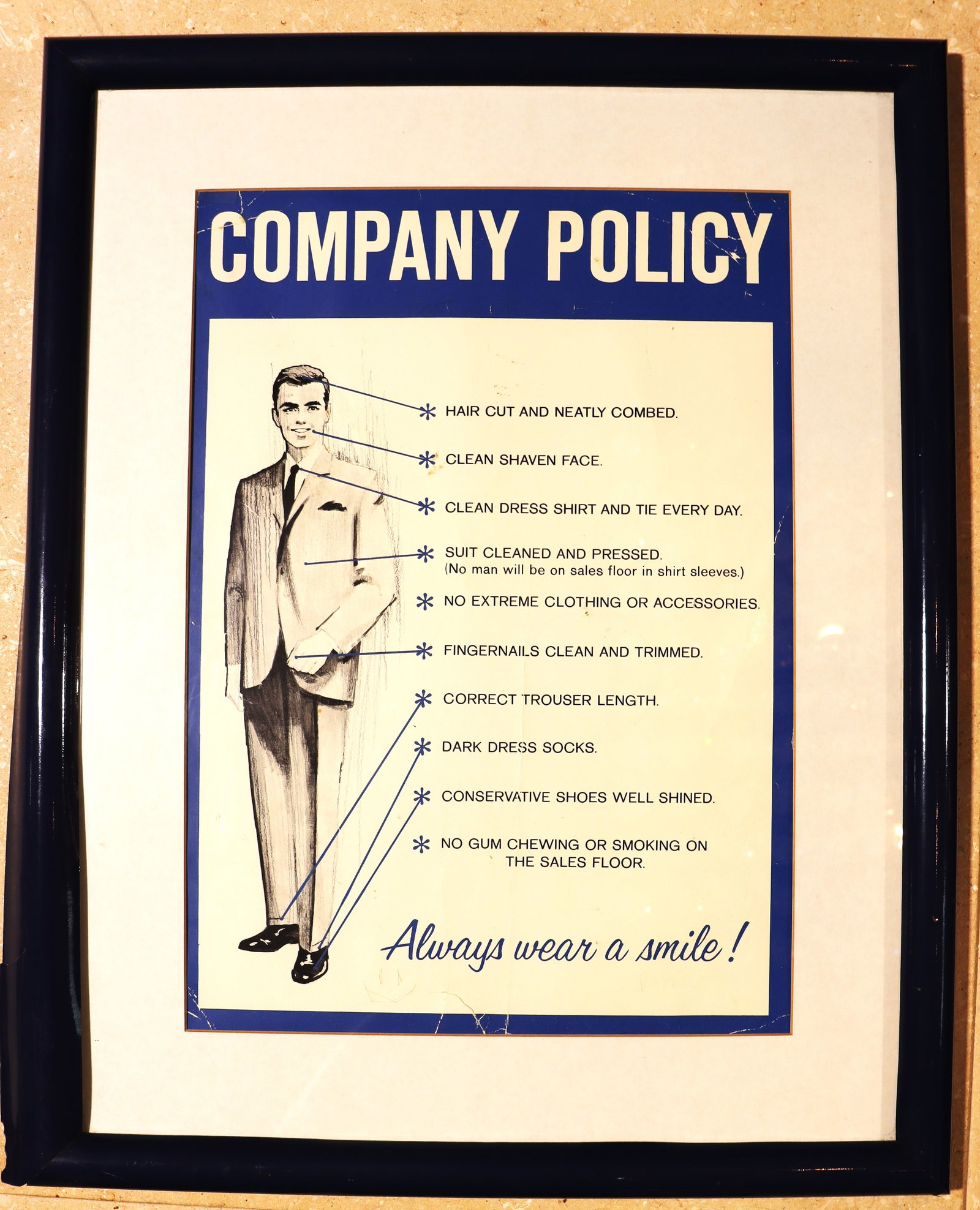 Framed "Company Policy" sign outlining a strict dress code for sales floor employees. The policy is: Framed "Company Policy" sign outlining a strict dress code for sales floor employees. The policy is highly prescriptive, emphasizing a professional and conservative