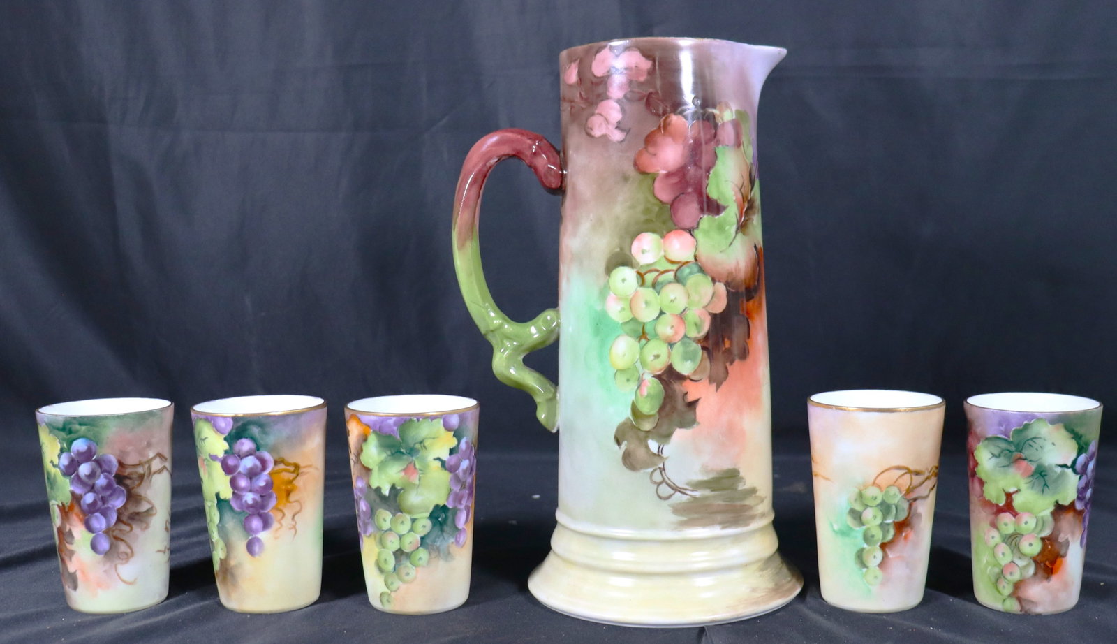 hand-painted porcelain tankard pitcher and matching mugs decorated with a cascading grape motif in: Hand-painted porcelain tankard pitcher and matching mugs decorated with a cascading grape motif in red, purple, and green on a soft green background. The Pitcher has specific maker's mark of 