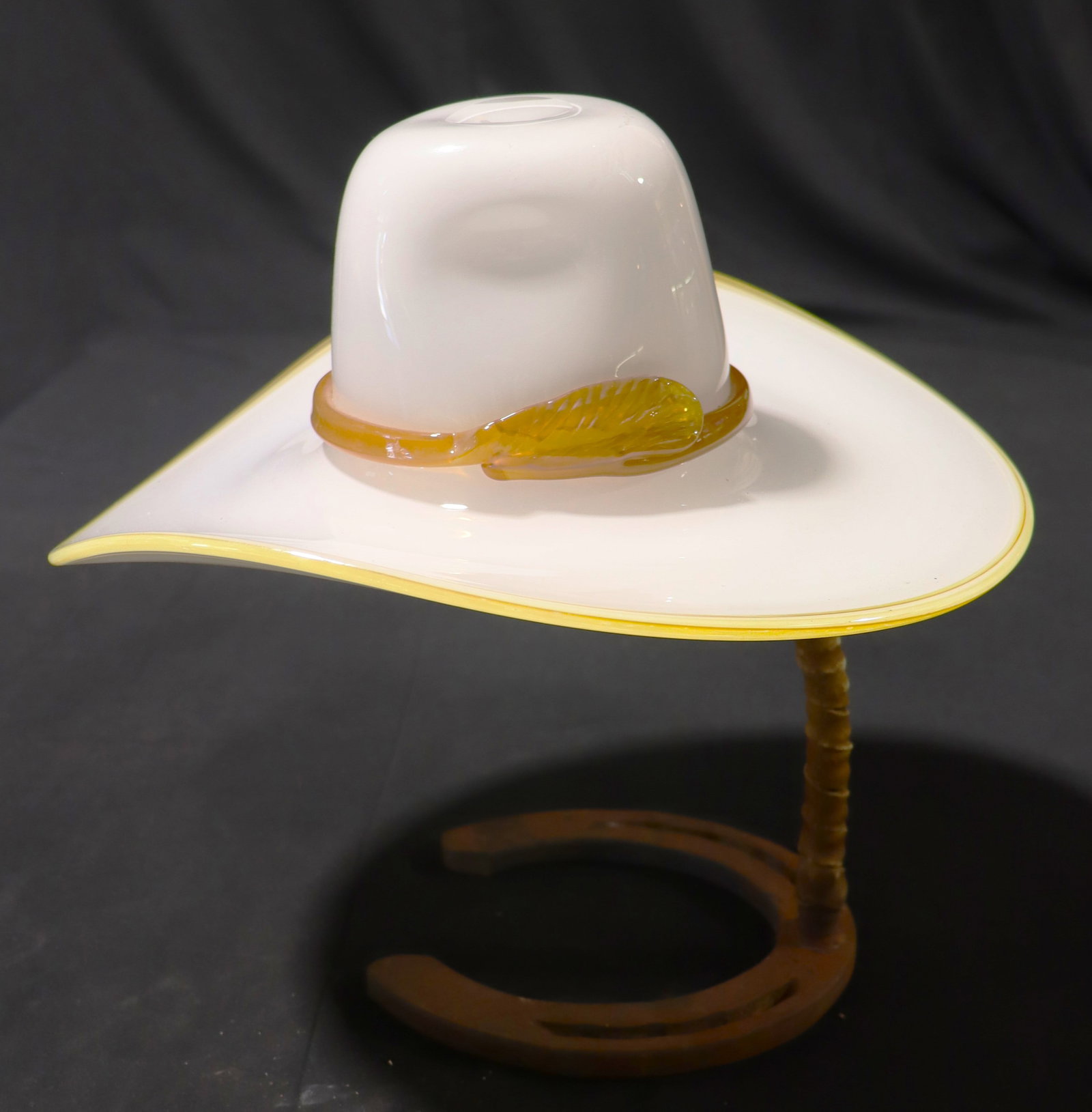 Vintage hand-blown art glass sculpture in the shape of a cowboy hat: Vintage hand-blown art glass sculpture in the shape of a cowboy hat, displayed on a decorative stand made from a real horseshoe and leather-wrapped post The piece is a western-style cowboy hat wi