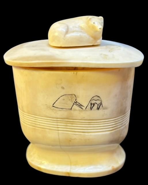 Alaskan Carved Walrus Tusk Lidded Dish with Scrimshaw: Alaskan hand carved walrus tusk lidded dish. Scrimshaw type design of two walrus heads on one side, and seals heads on other. Lid has an applied carved fox figure. Measures 3 5/8". Ca early 19th centu