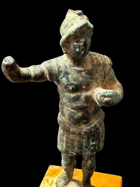 Bronze Roman Solider Figure on Stone Pedestal: Bronze soldier wearing knee-length tunic and Phrygian cap, poses with one arm extended - a common pose for Roman deities on a stromatolite rock base. Measures 5.5" high by 1.75" wide. Age unknown.
