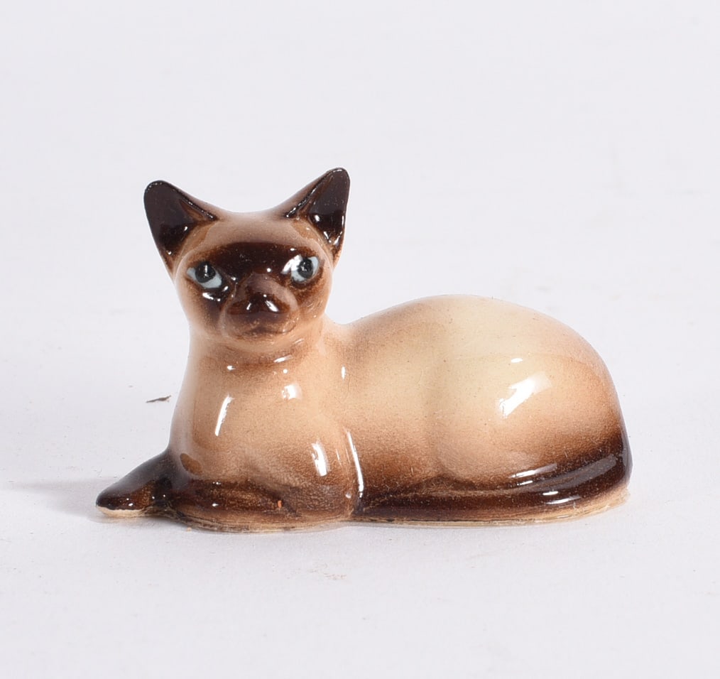 Reclining Siamese Cat Figurine: Vintage glazed ceramic Siamese cat figurine modeled reclining with dark facial mask and expressive blue eyes.