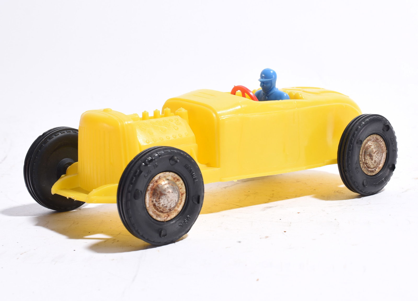 Driver Figure Roadster Race Car: Open-wheel racing roadster with yellow body, blue driver figure, and red steering wheel. Features exposed engine detail and black wheels. Vintage plastic construction with light wear consistent with u