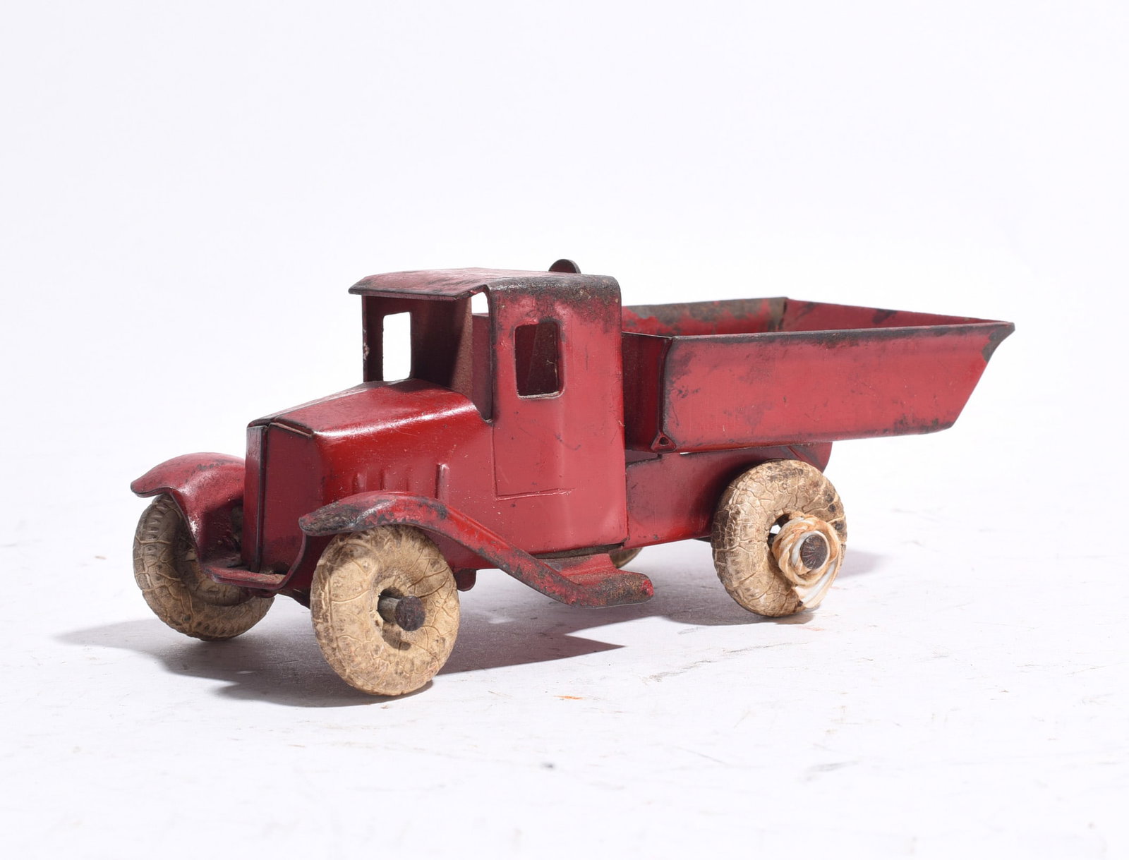 Early Pressed Steel Dump Truck: Early pressed steel dump truck with red painted body and dump bed. Retains original composition-style wheels showing significant age-related deterioration. Displays oxidation, paint wear, and surface 