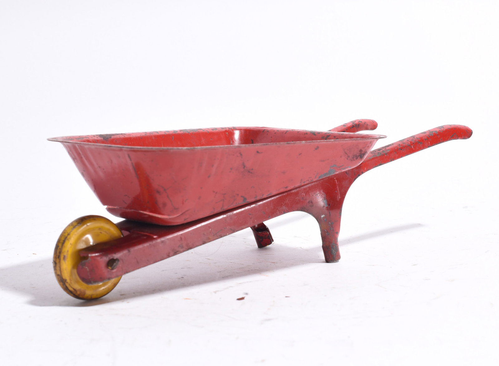 Toy Wheelbarrow: Child’s toy wheelbarrow constructed of painted metal with red body and yellow wheel. Displays wear, scratches, and paint losses consistent with use.