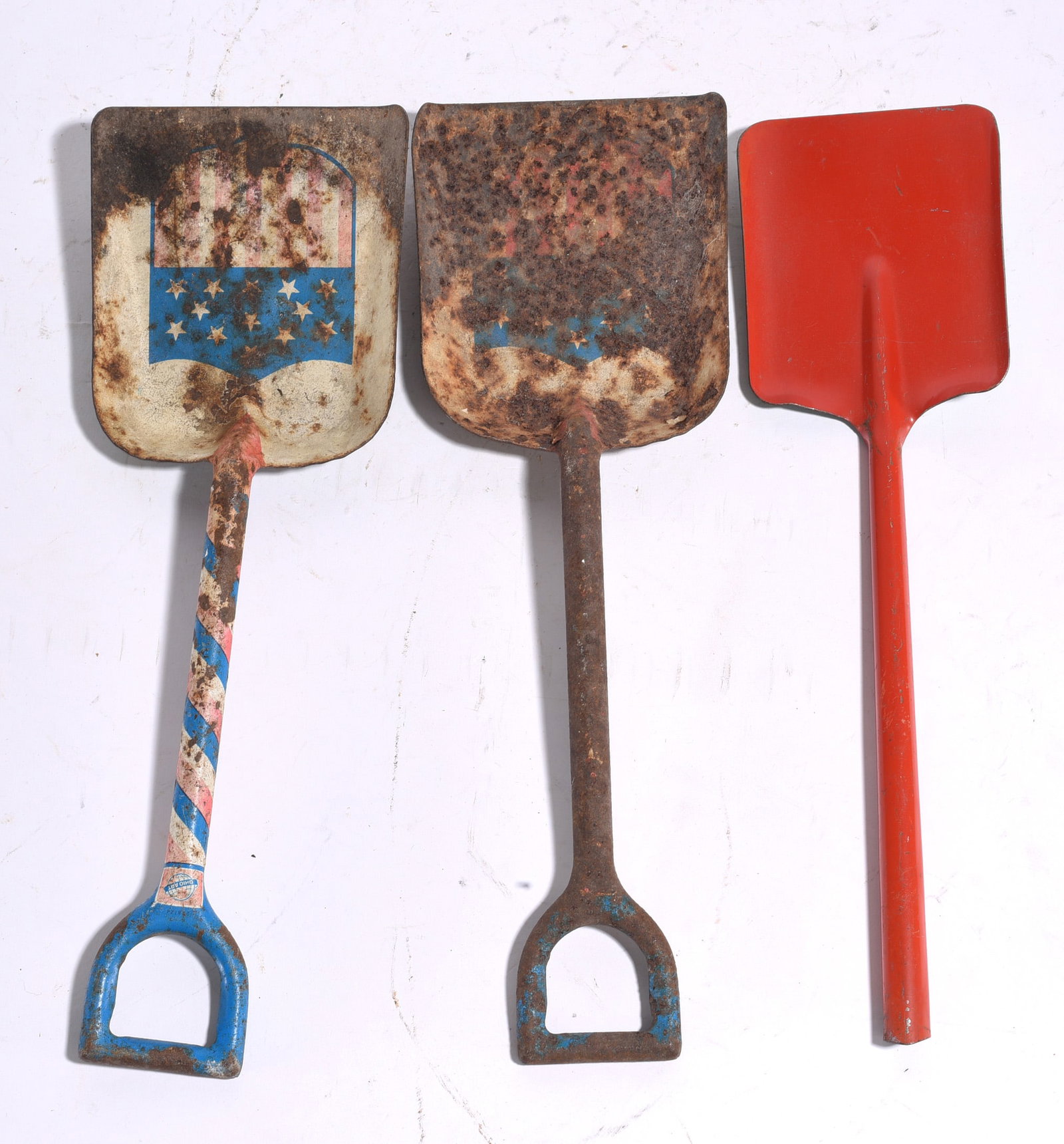 Patriotic Child's Sand Shovel Group: Lot of three vintage toy sand shovels including two metal examples with patriotic shield decoration and one red-painted shovel. Showing play wear, oxidation, and surface rust consistent with age and