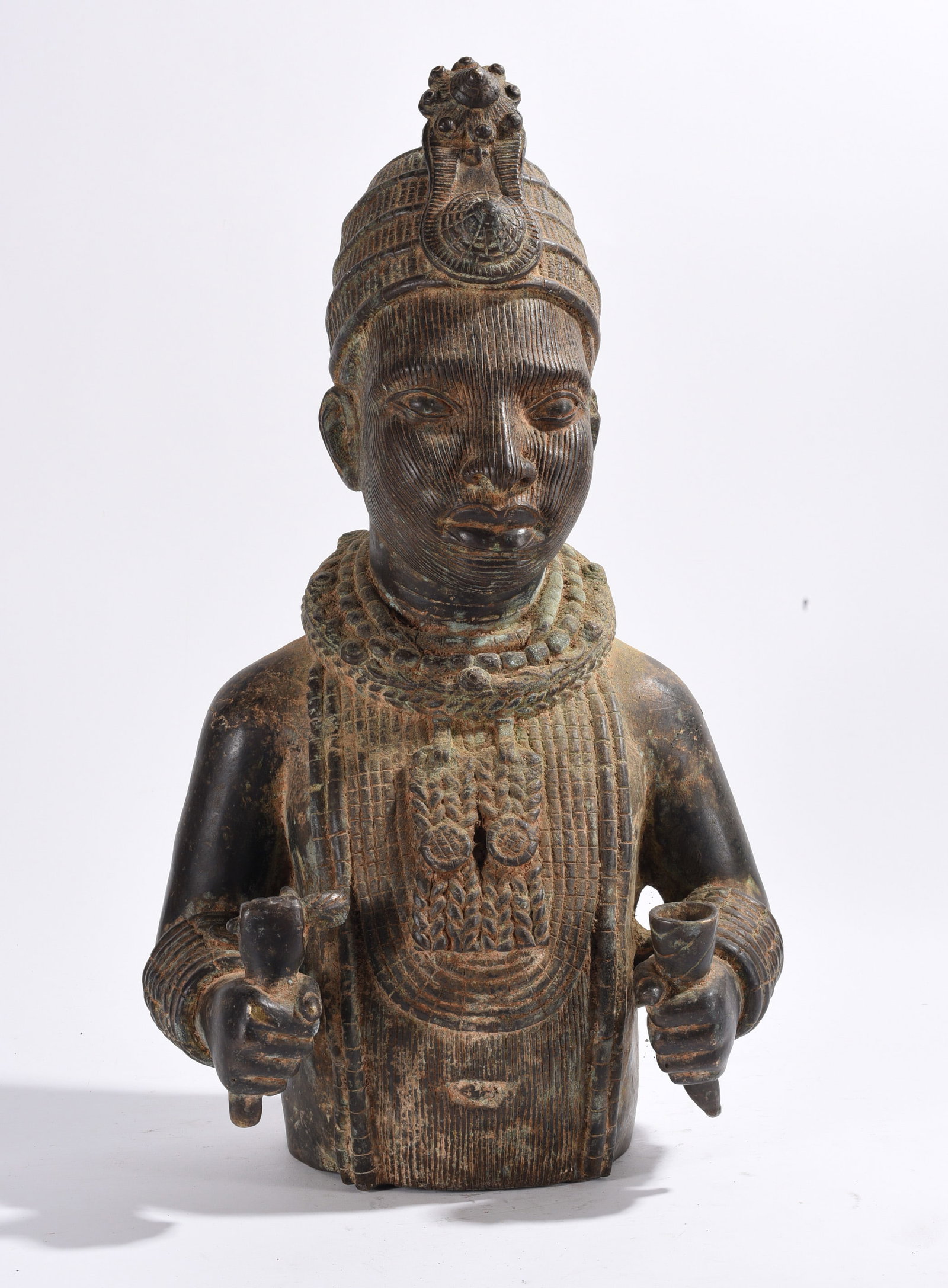 Benin Style Bronze Royal Bust Figure: Cast bronze royal bust depicting a dignitary holding ceremonial objects. Finely incised robes, jewelry, and headdress details demonstrate skilled lost-wax casting techniques. Exhibits an aged green-br