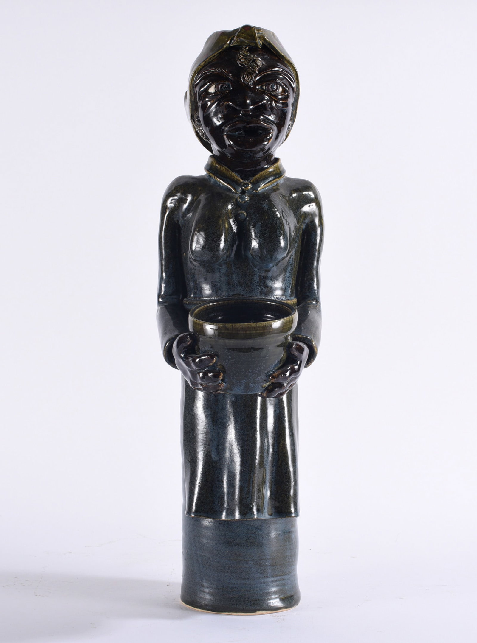 Lyn Craven Large Folk Pottery Figural: 21" Tall folk pottery figural depicting a woman holding a vessel at her waist, finished in a deep dark glaze with modeled facial features, hands, clothing and head covering.