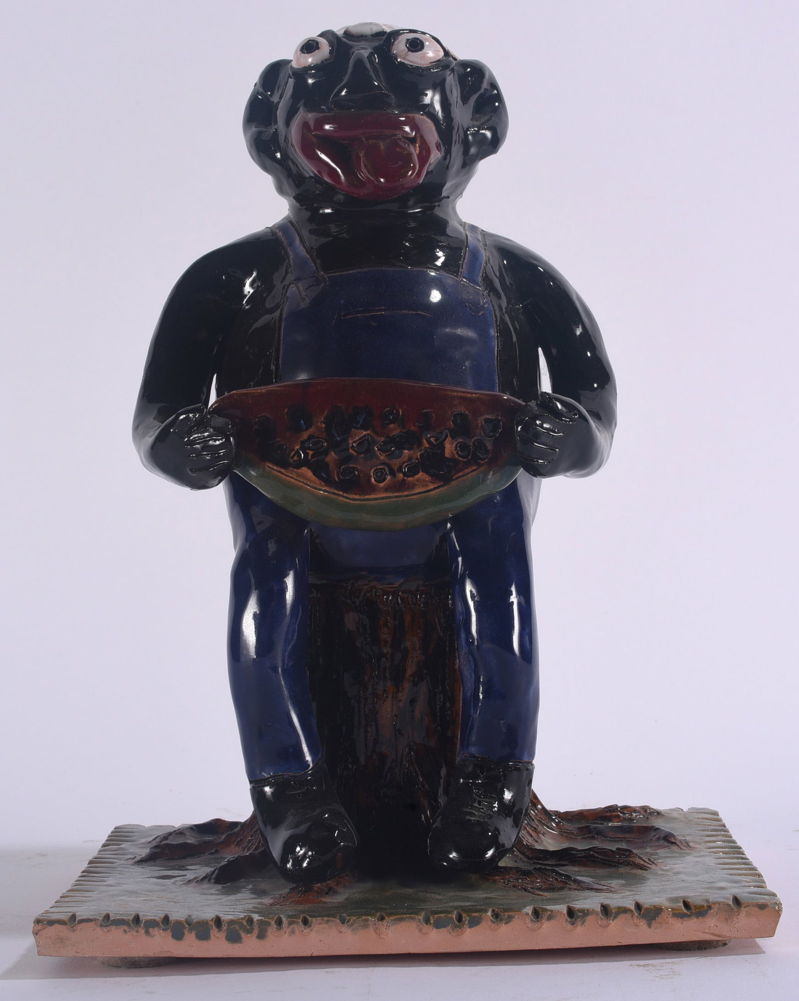 Claude Miller Folk Pottery Figural: 12" Large figural folk pottery sculpture by North Carolina potter Claude Miller, depicting a seated Black male figure holding a watermelon. Boldly modeled with exaggerated facial features, applied eye