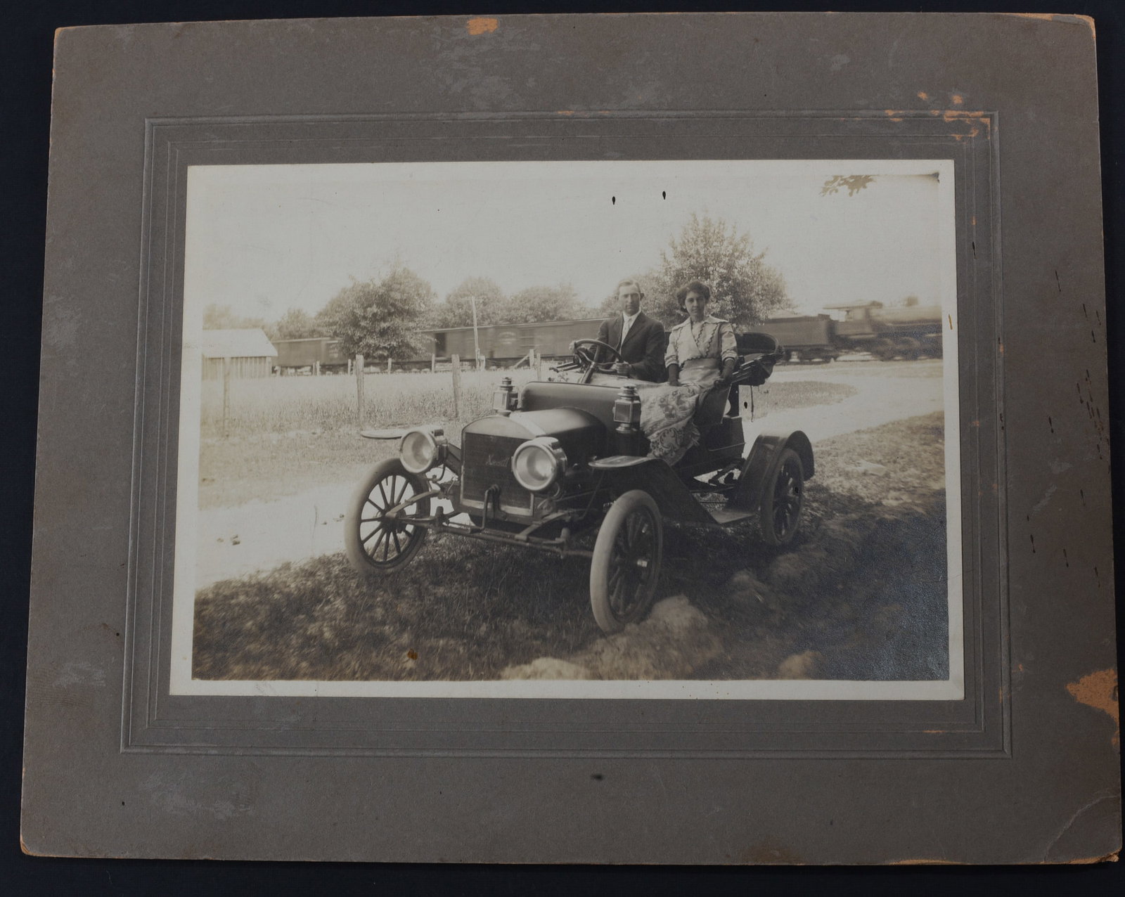 1912 Automobile Photograph: Albumen photograph 7”x5” of a couple in a early automobile. Id on verso of period paper mount “Jno B. Lane & Miss Lally Ziegler 5/20/12” . With R.L. Grimm, Artist stamp. Good
